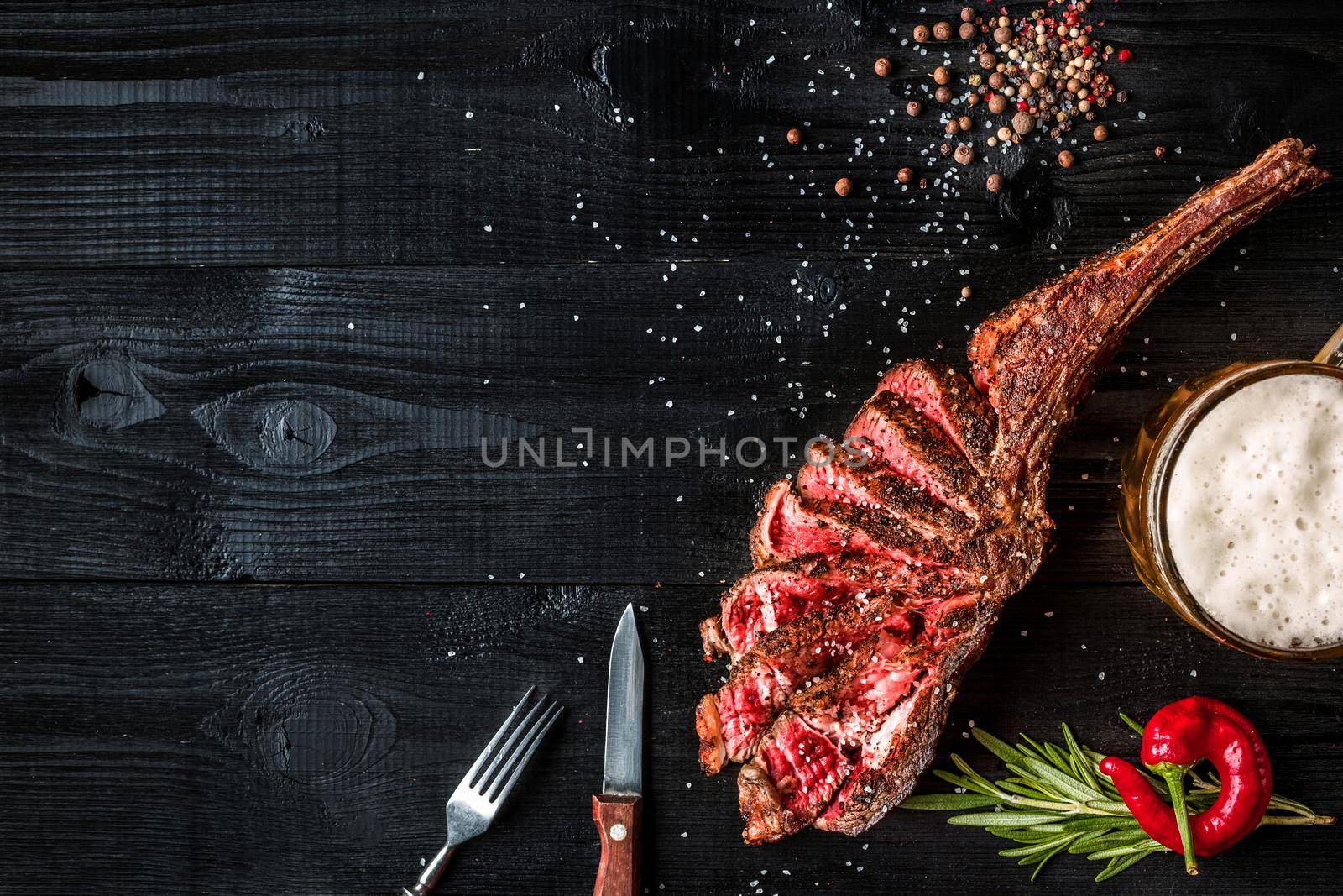 Barbecue dry aged rib of beef with spice, vegetables and a glass of light beer close-up on black wooden background. Top view. Copy space. Still life. Flat lay