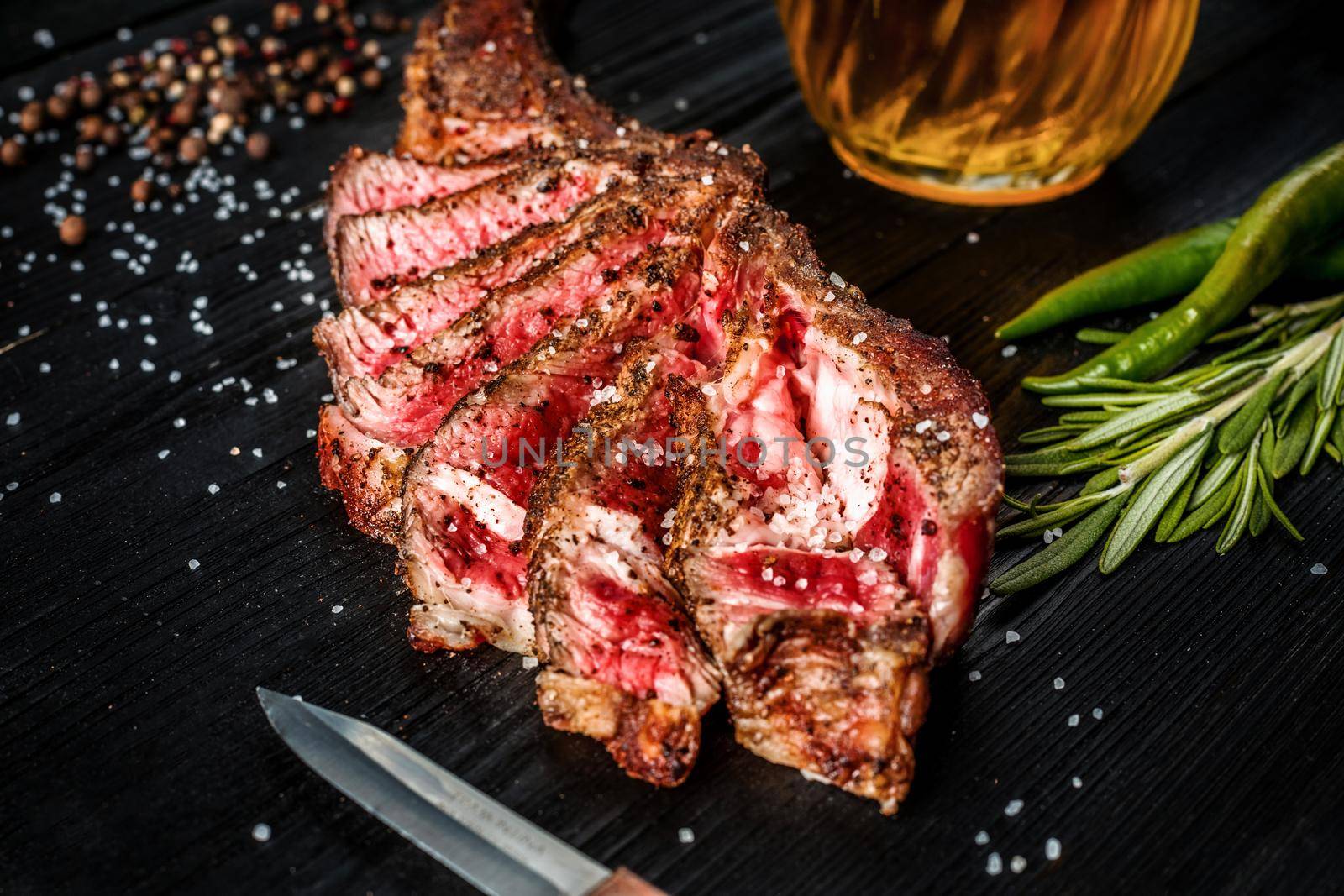 Barbecue dry aged rib of beef with spice, vegetables and a glass of light beer close-up on black wooden background. Top view. Copy space. Still life. Flat lay