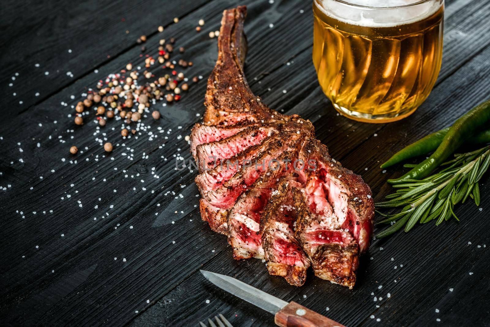 Barbecue dry aged rib of beef with spice, vegetables and a glass of light beer close-up on black wooden background. Top view. Copy space. Still life. Flat lay