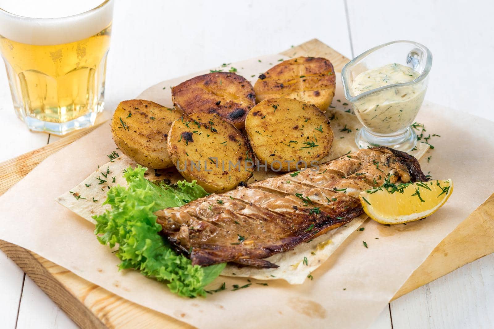 Fish fillet, fried potatoes with greens, lemon and sauce on a wooden board. A glass of light beer and a ready fish dish on a white wooden table