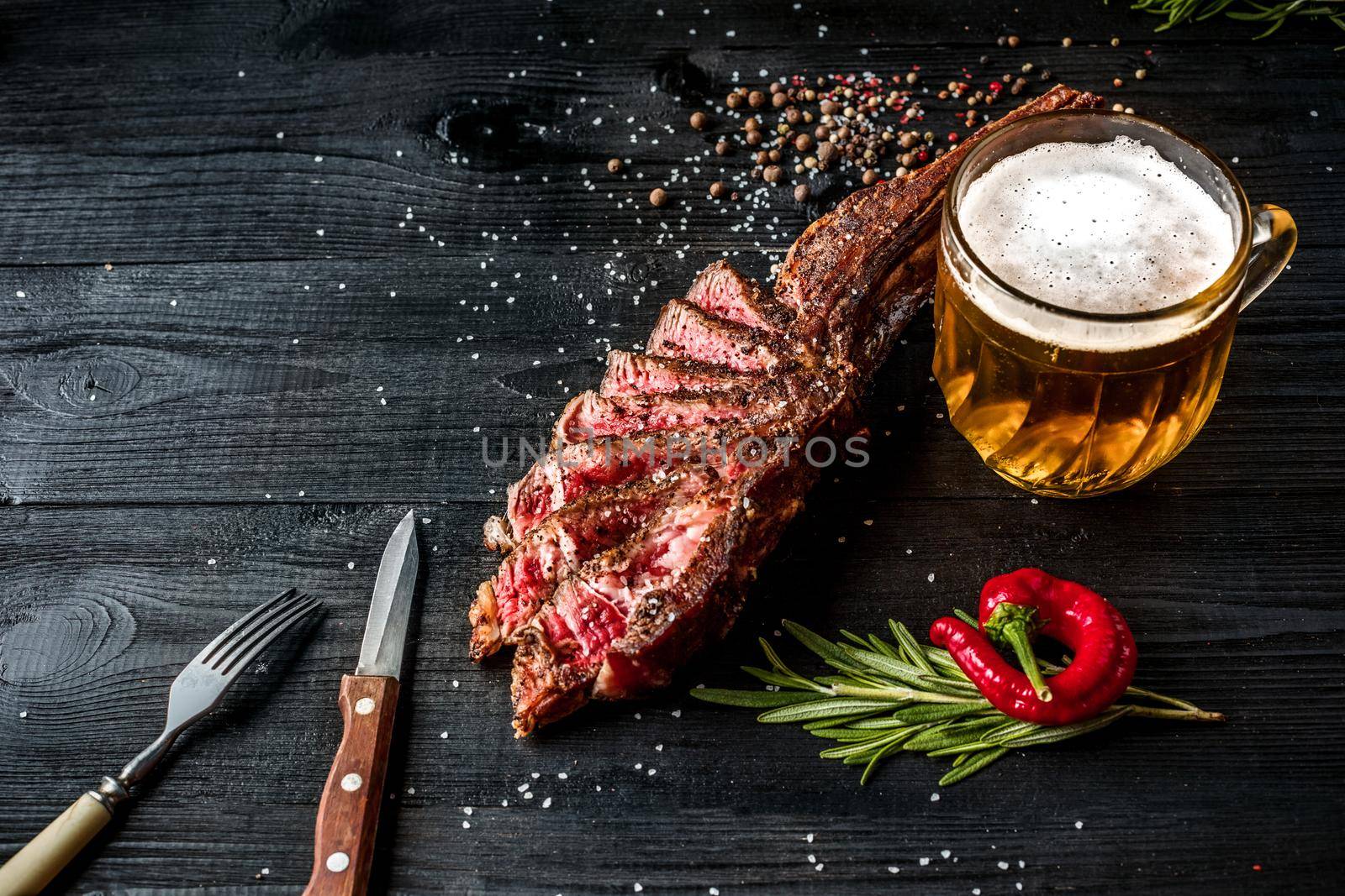 Barbecue dry aged rib of beef with spice, vegetables and a glass of light beer close-up on black wooden background by nazarovsergey