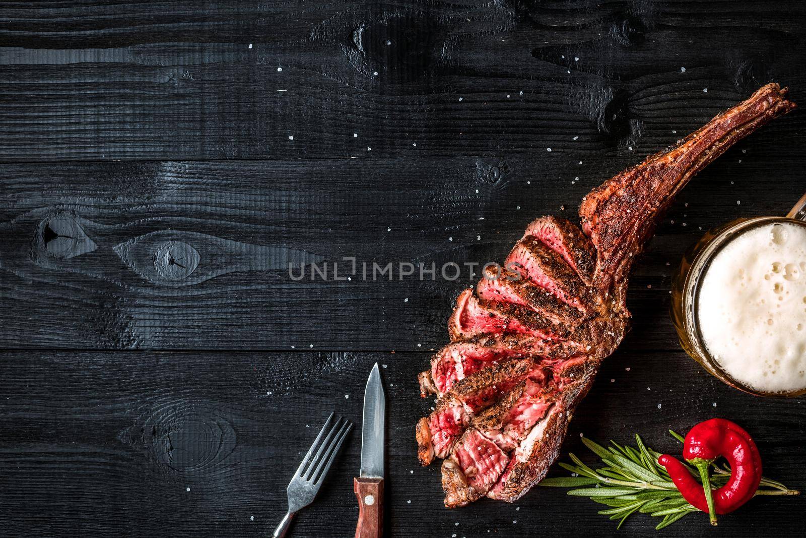 Barbecue dry aged rib of beef with spice, vegetables and a glass of light beer close-up on black wooden background. Top view. Copy space. Still life. Flat lay