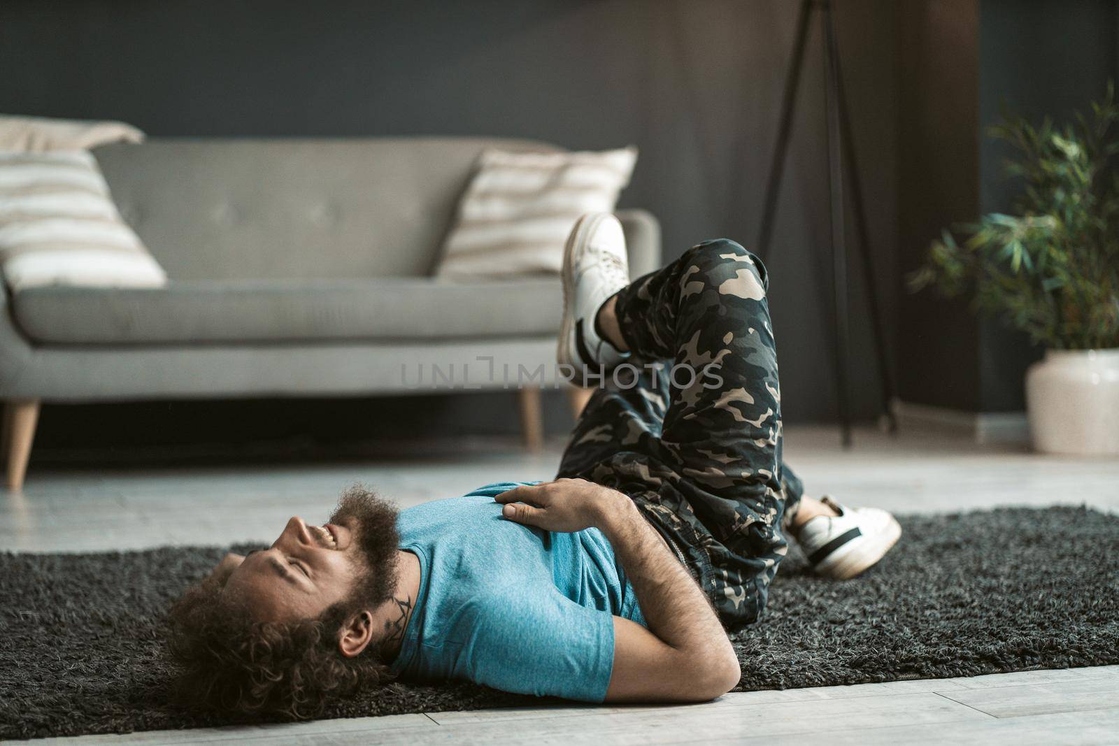 Handsome Slender Guy is having Fun on the Carpet on the Floor in the Middle of the Room. Man in a Blue T-shirt and Military Pants has a Rest on the Carpet. Gray Sofa Background. Copy space . Close-up . High quality photo
