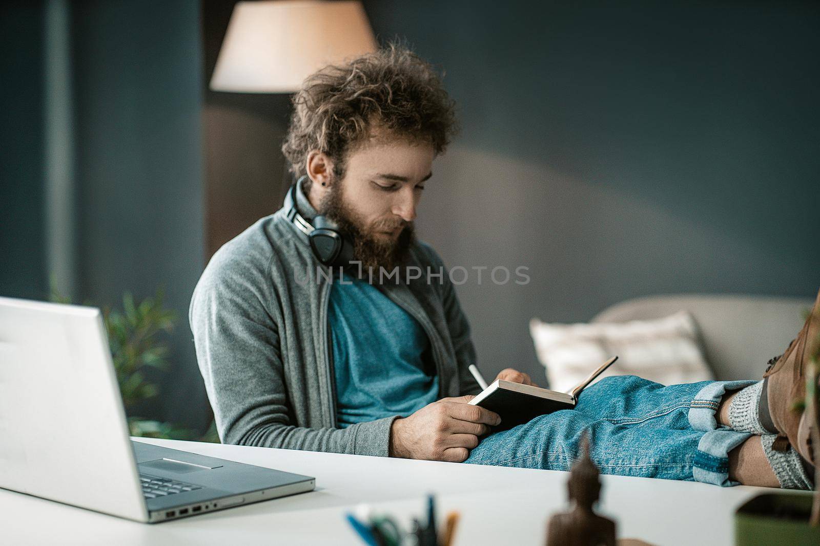 Student Listens to an Online Lecture and Makes Notes in a Notebook. a Man Writes Important Information in a Diary. Headphones Around his Neck. Feet on the Table. Close-up. High quality photo