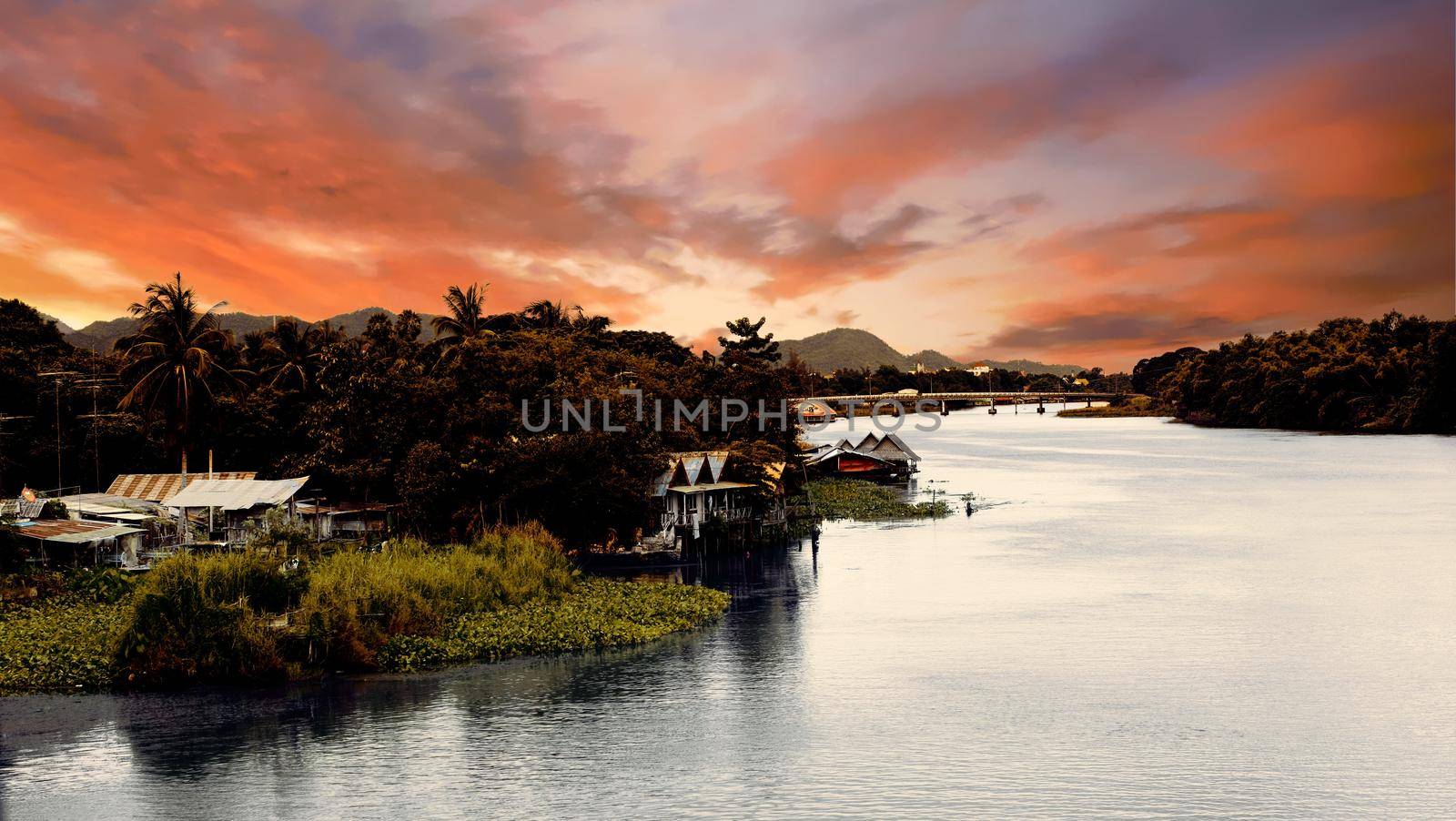 Dark contrast sunset in tropics on big river with village, bridge and mountains on background. The Bridge on the River Kwai, Thailand 5th of july 2009. High quality photo