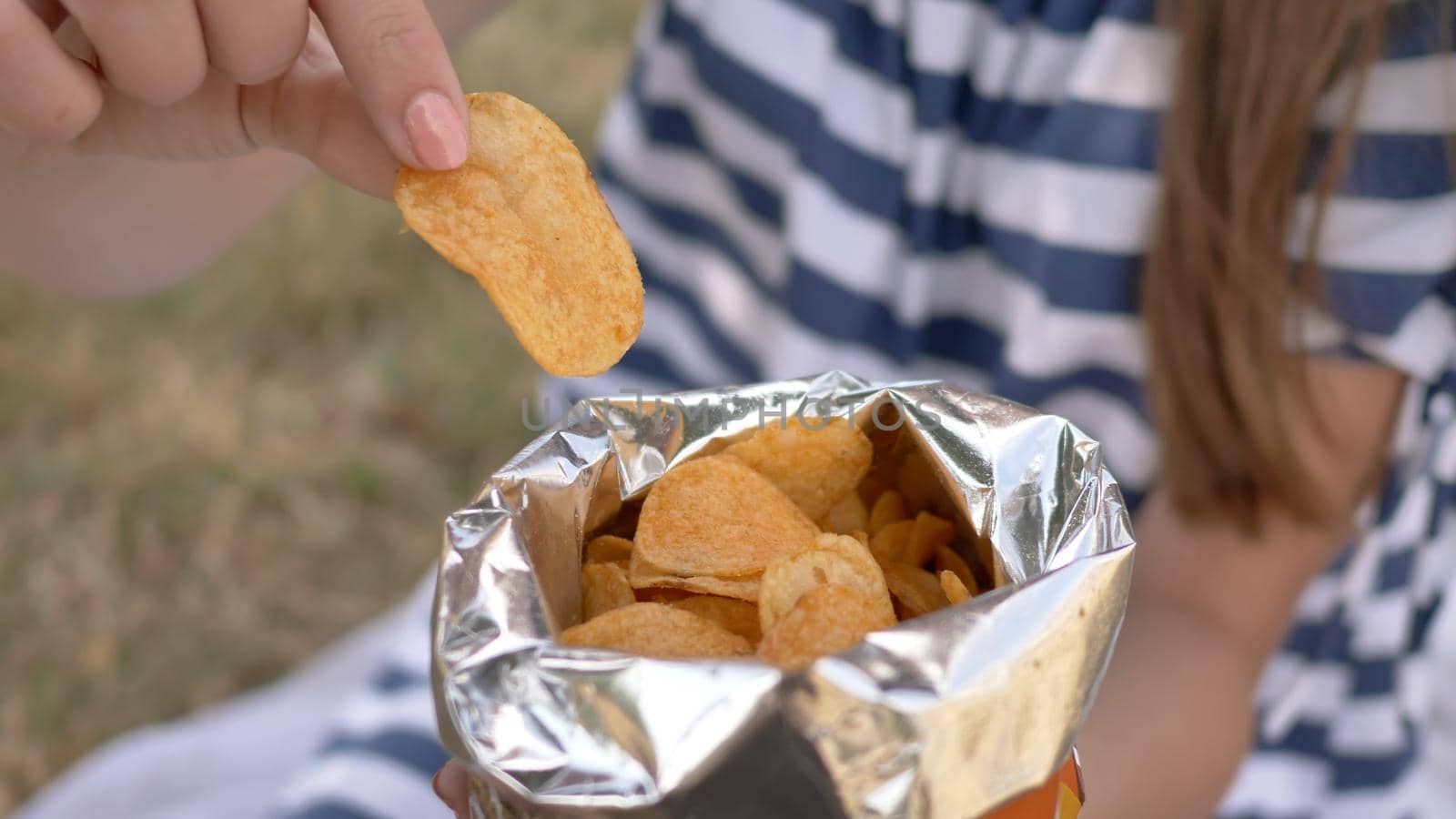 The girl eats chips in the city park in the summer. The girl eats chips in the city park in the summer. Package with chips close-up. by DovidPro