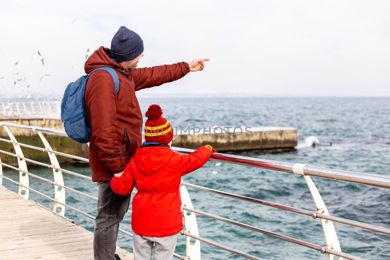 Father and son standing on a pier looking at the sea in winter. Man pointing at something ahead. Travel lifestyle exterior