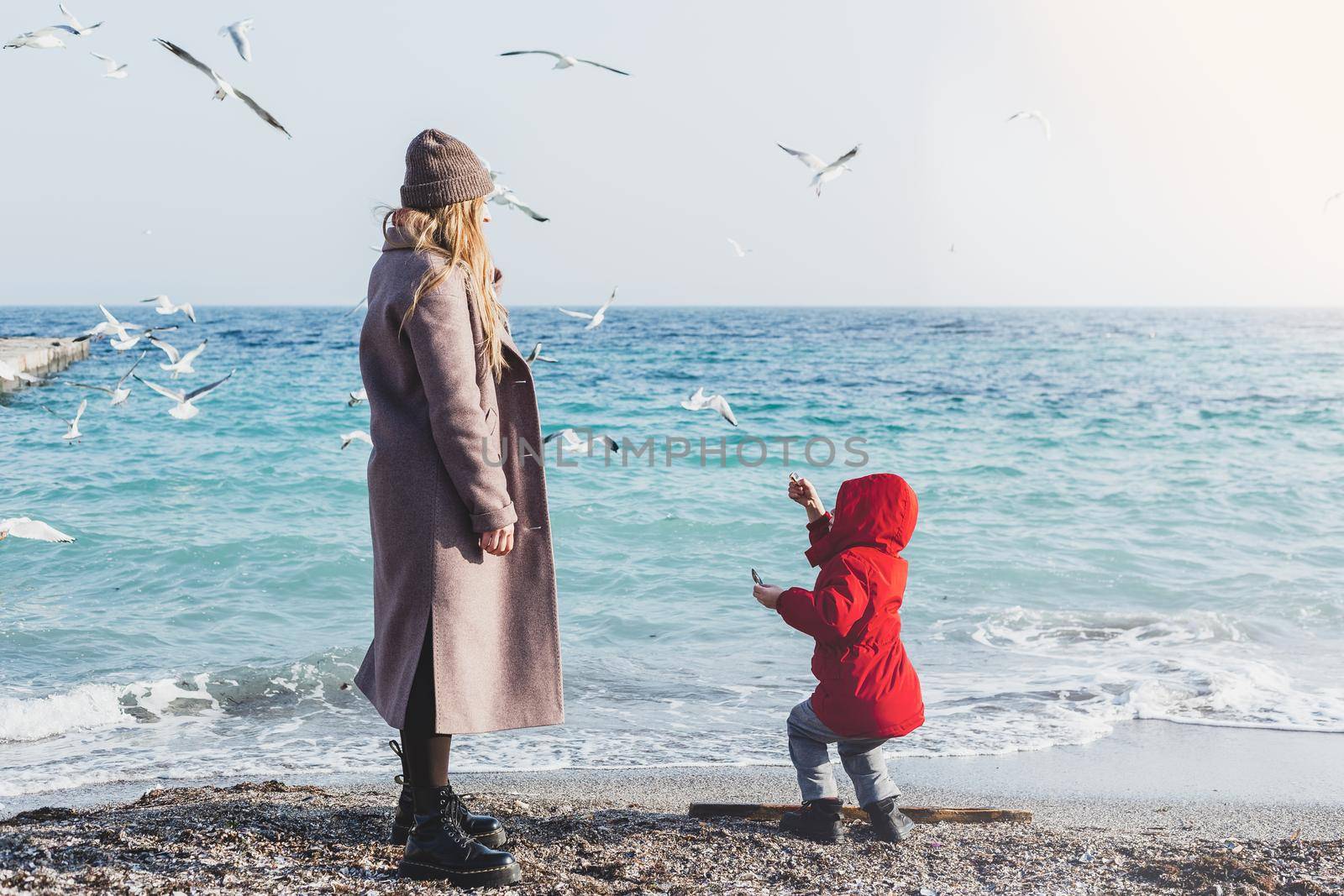 Mother and son walk to the seaside. Birds flying above the water. Cold season vacations