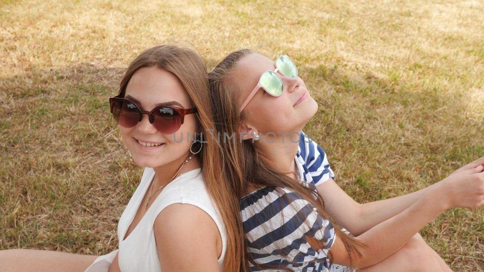Cheerful female friends in sunglasses are sitting on the grass in the city. by DovidPro