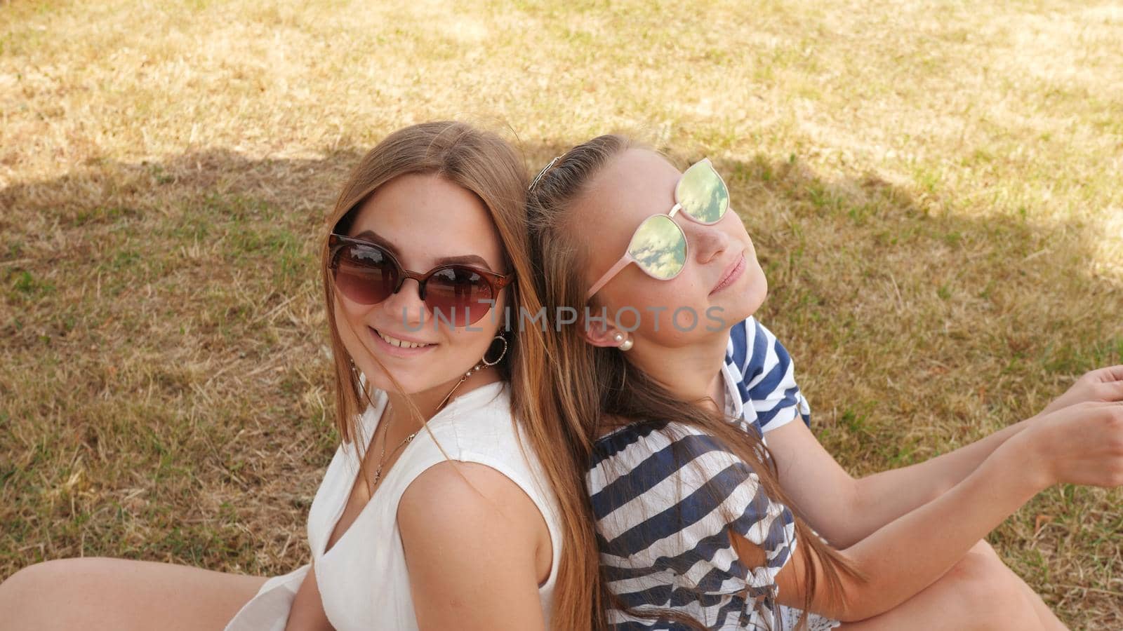 Cheerful female friends in sunglasses are sitting on the grass in the city. by DovidPro