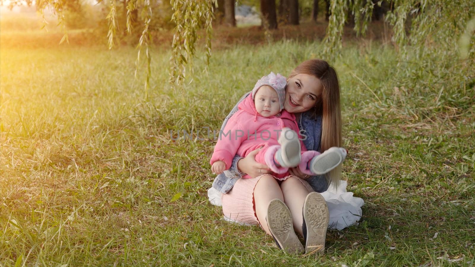 Happy young mother with her baby is sitting on the grass in the park in the autumn evening. by DovidPro