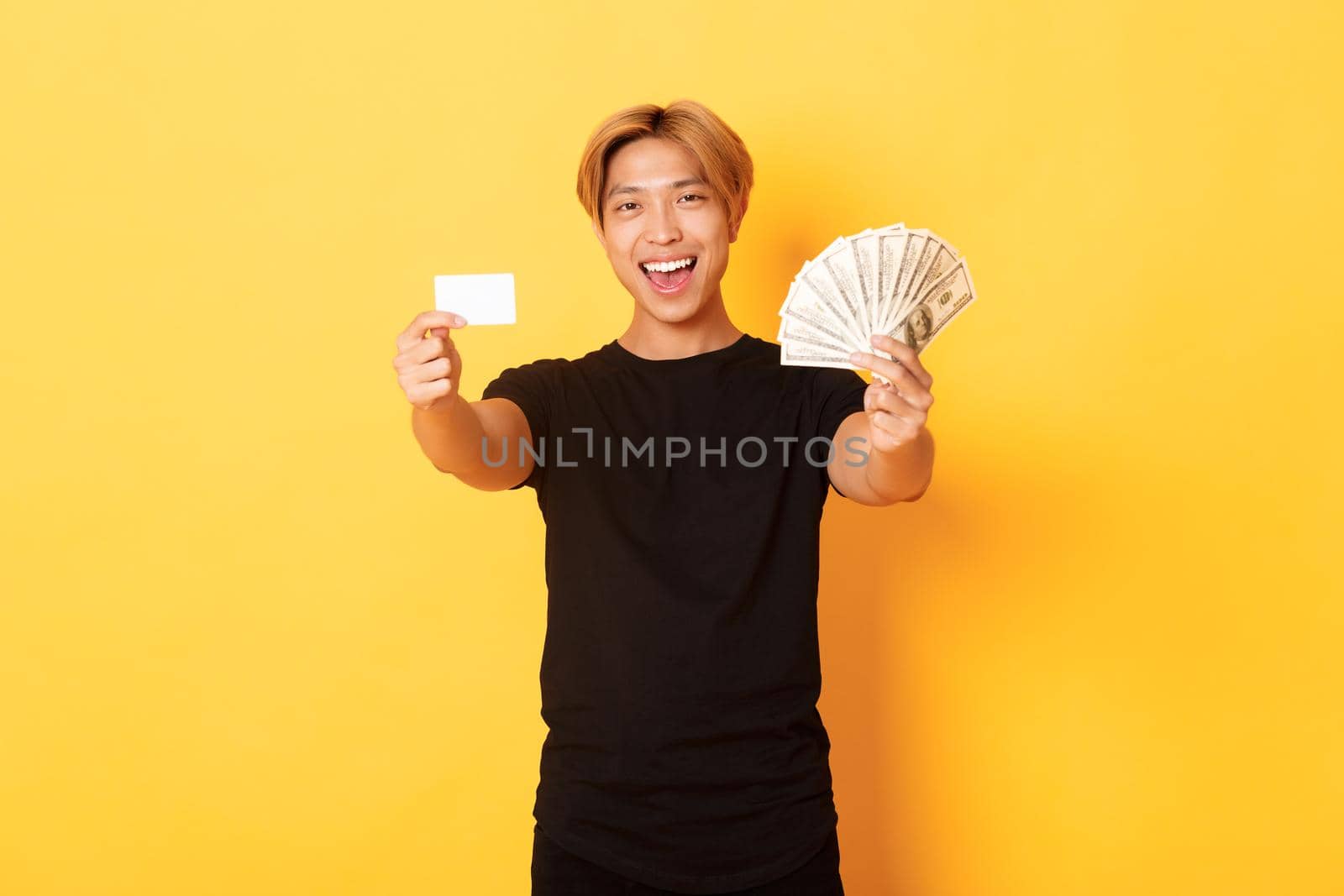 Happy good-looking asian guy in black casual clothes, showing money and credit card, smiling sassy, yellow background by Benzoix