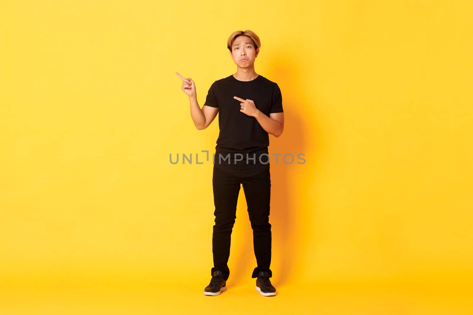 Full-length of sad handsome korean guy with blond hair making upset grimace, pointing fingers upper left corner, showing logo with regret, standing yellow background by Benzoix