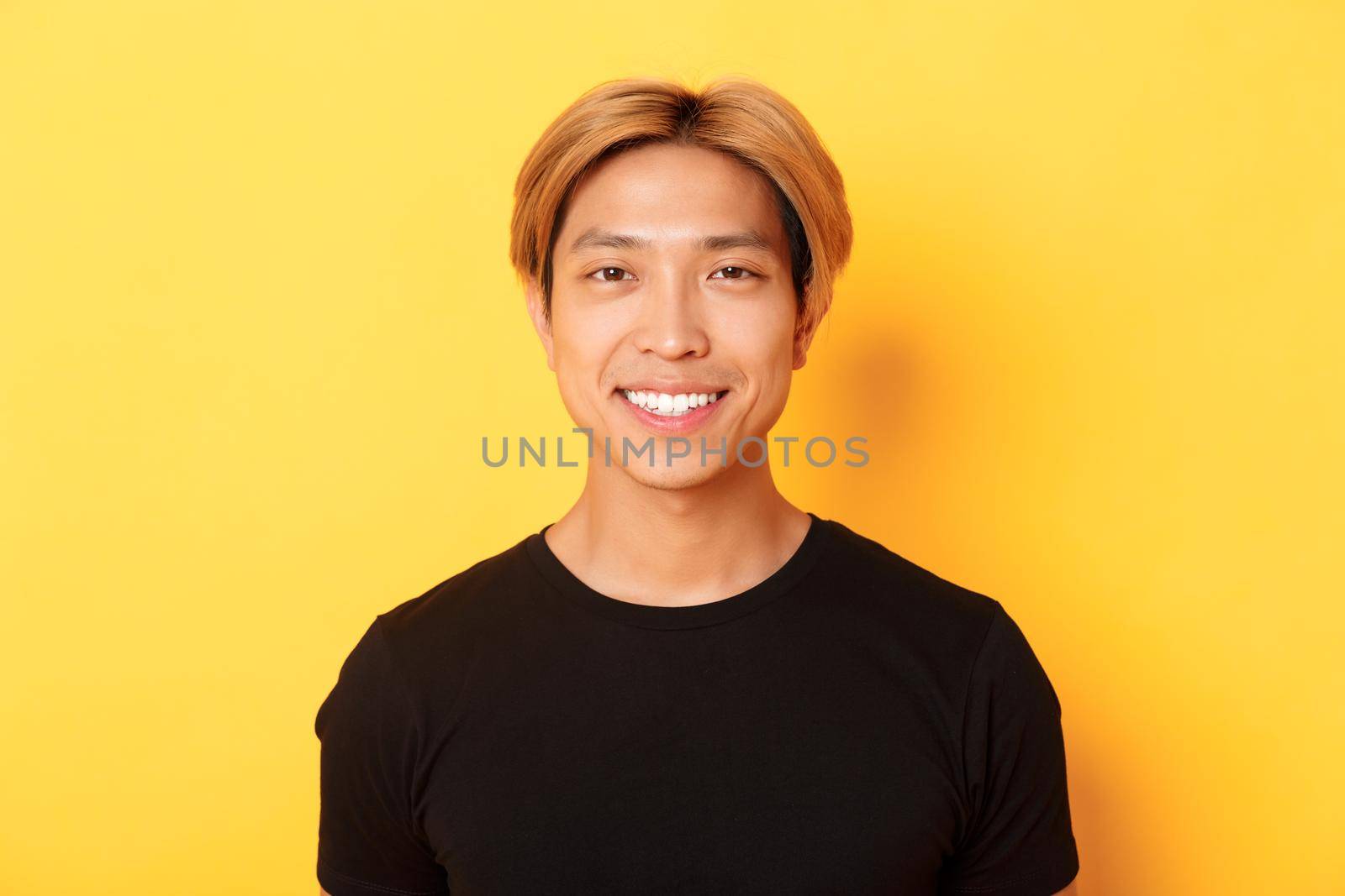 Close-up of handsome blond asian guy in black t-shirt, smiling happy at camera, standing over yellow background by Benzoix