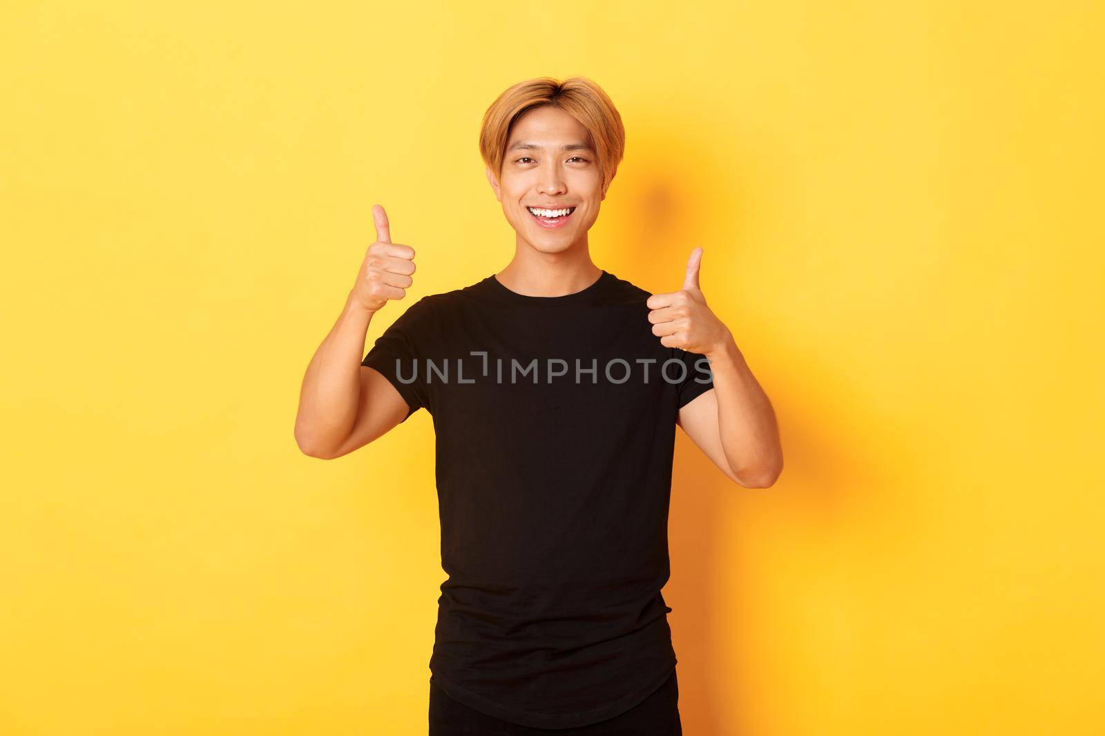 Portrait of satisfied asian man smiling and showing thumbs-up over yellow background by Benzoix