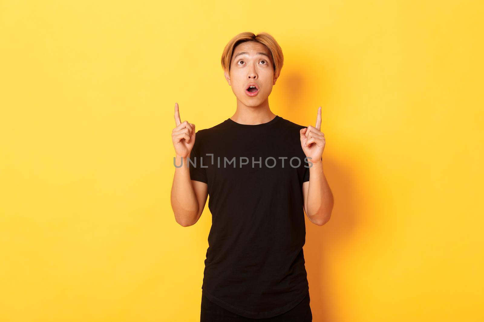 Portrait of curious and amazed asian guy with blond hair, wearing black t-shirt, looking and pointing fingers up astonished, yellow background by Benzoix