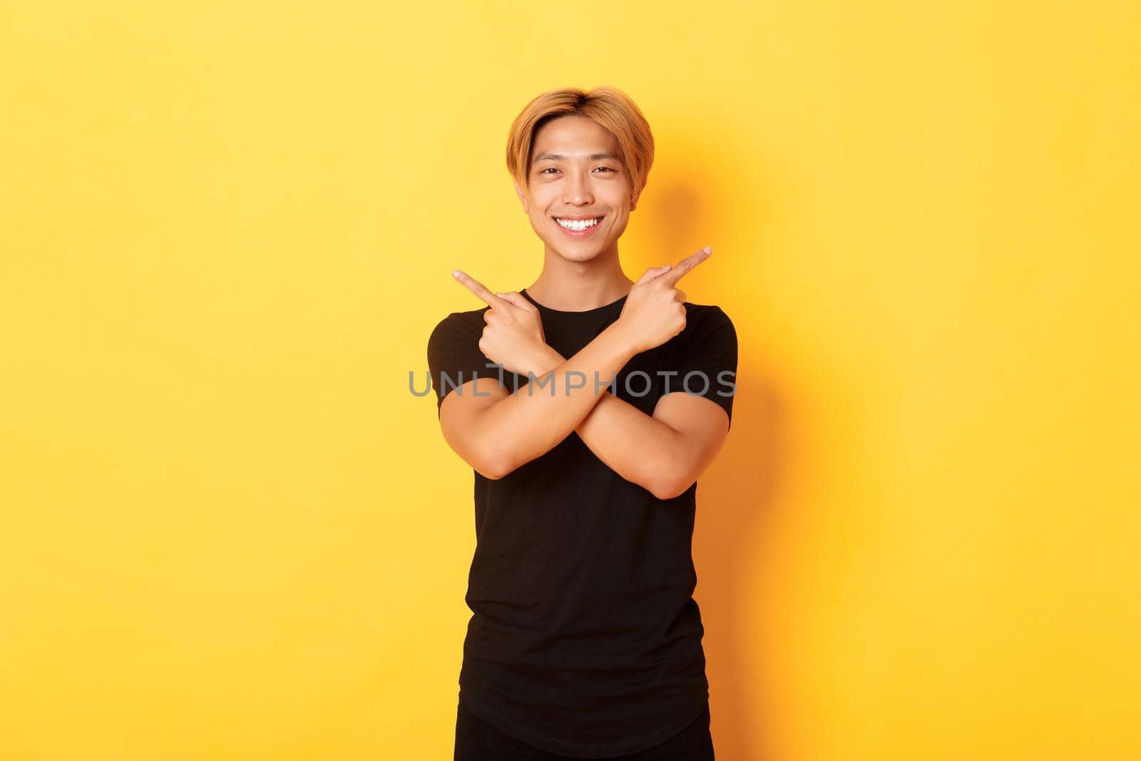 Portrait of handsome determined blond asian guy, student pointing fingers sideways, showing two variants, standing yellow background by Benzoix
