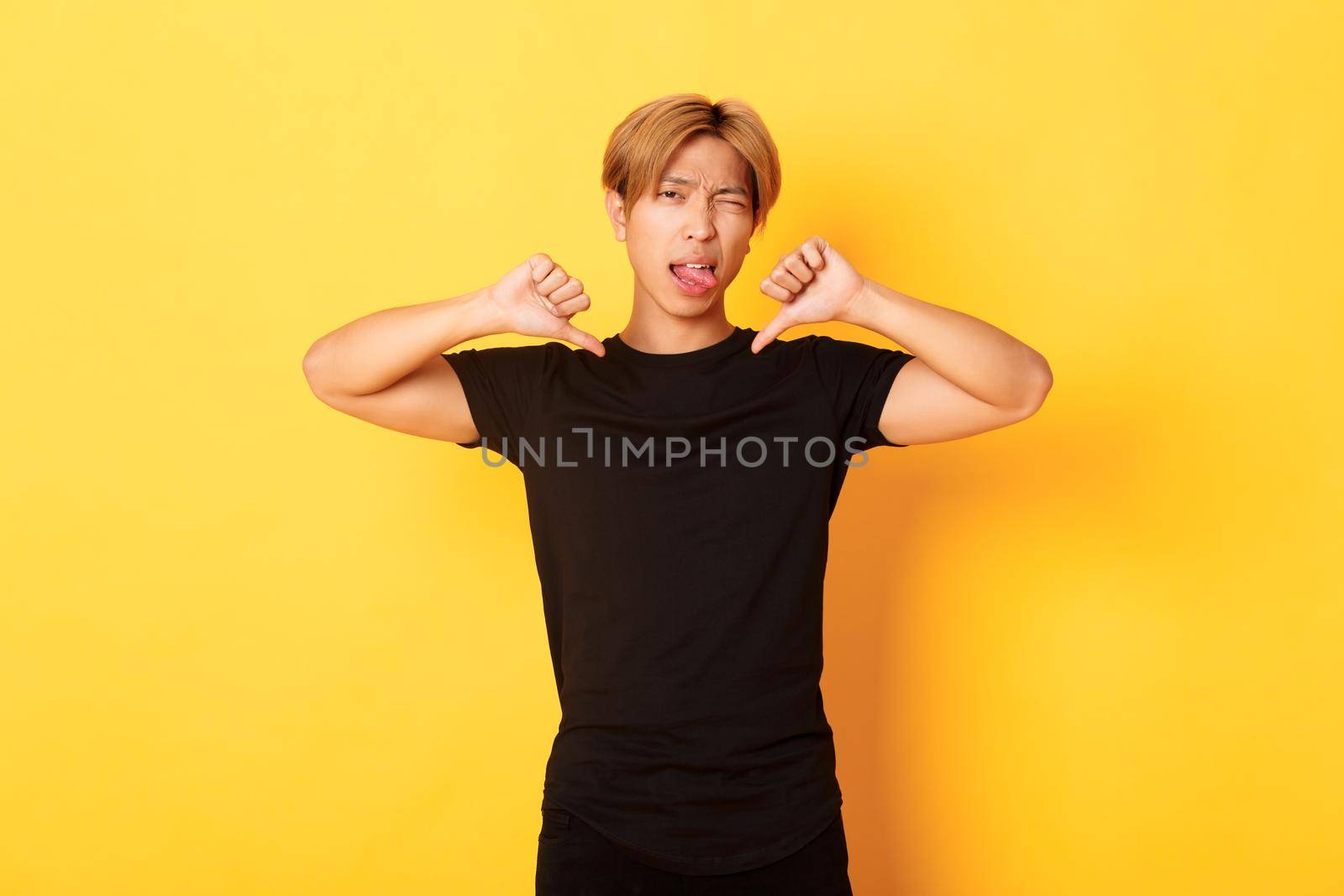 Portrait of sassy asian handsome guy showing thumbs-down and stick tongue disappointed, judge something bad, yellow background.