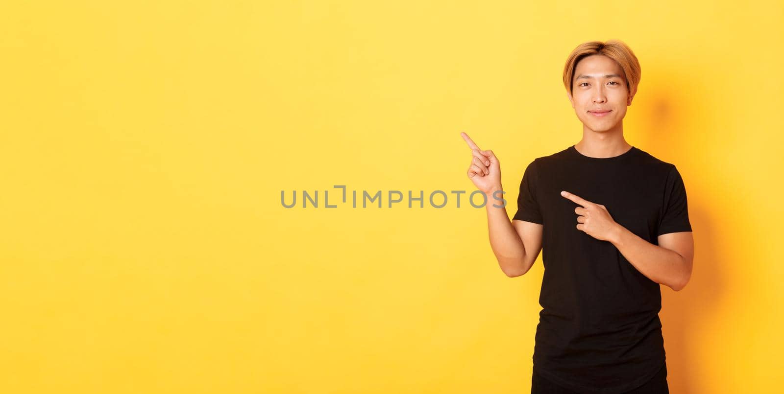 Portrait of smiling handsome asian man in black t-shirt pointing fingers upper left corner, showing logo or banner, yellow background.