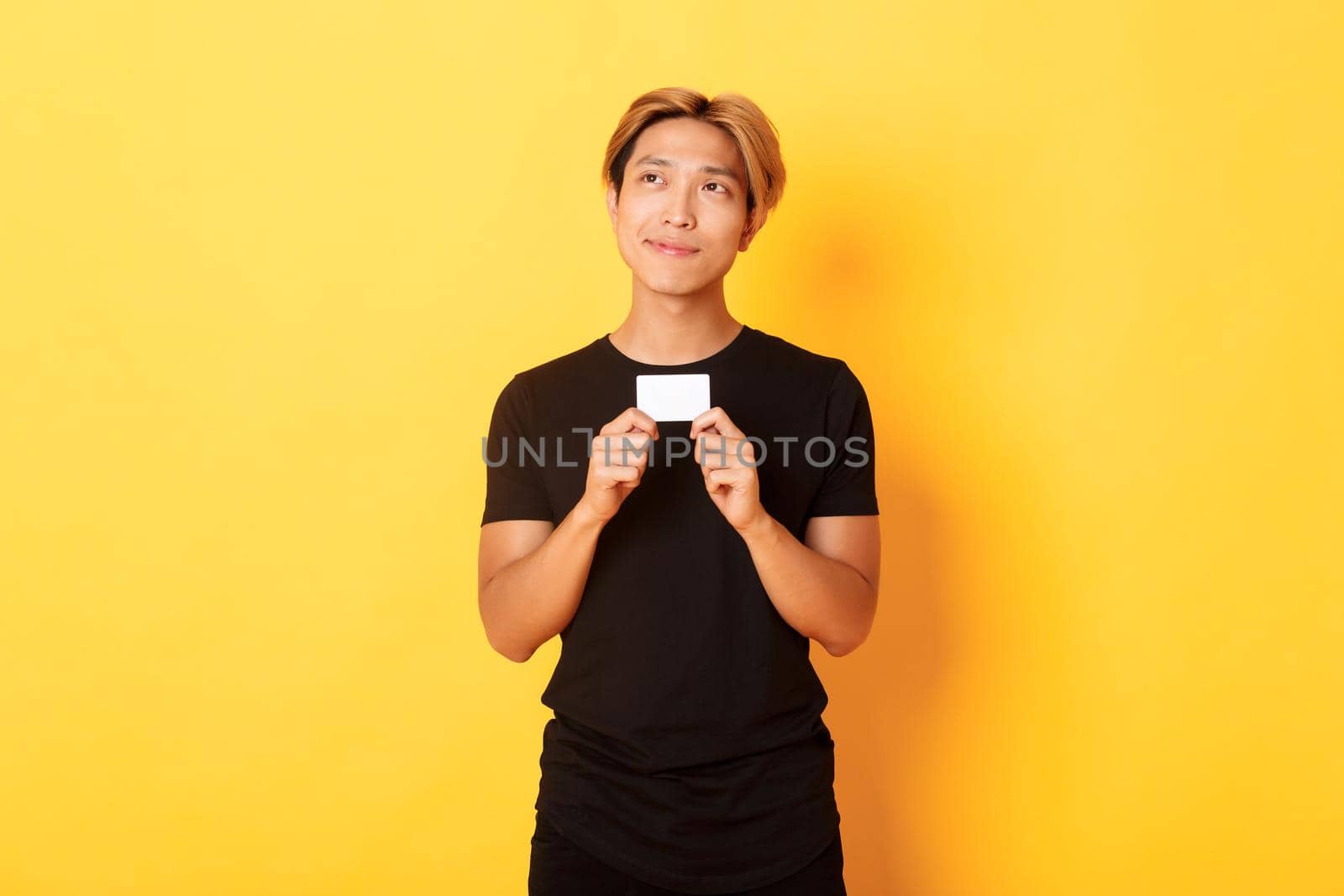 Thoughtful smiling asian guy thinking while showing credit card, looking upper left corner dreamy, yellow background by Benzoix