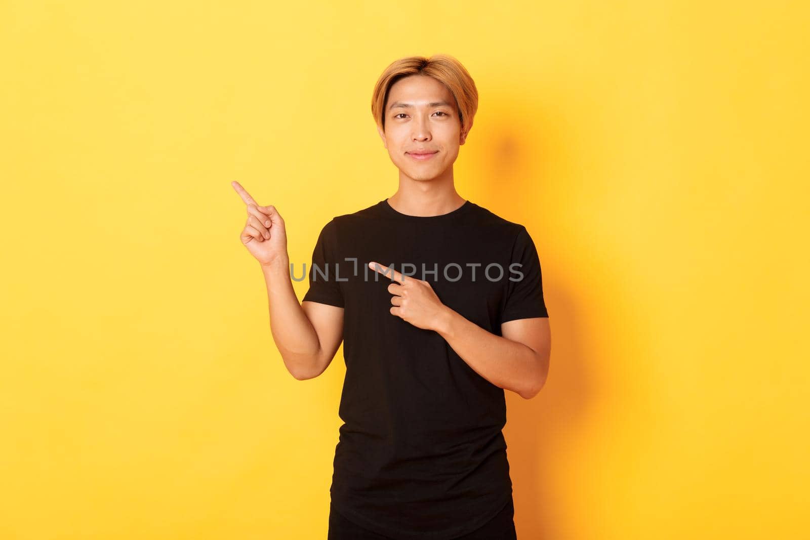 Portrait of smiling handsome asian man in black t-shirt pointing fingers upper left corner, showing logo or banner, yellow background.