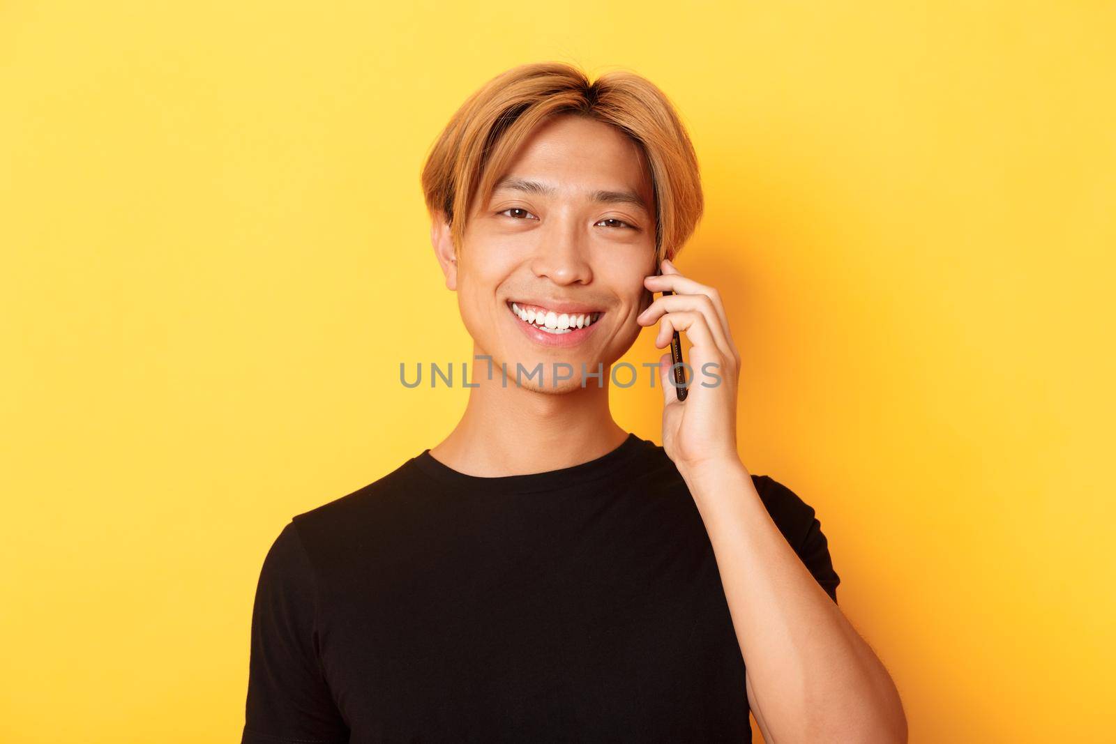 Close-up of handsome stylish korean guy smiling and talking on mobile phone, standing over yellow background.