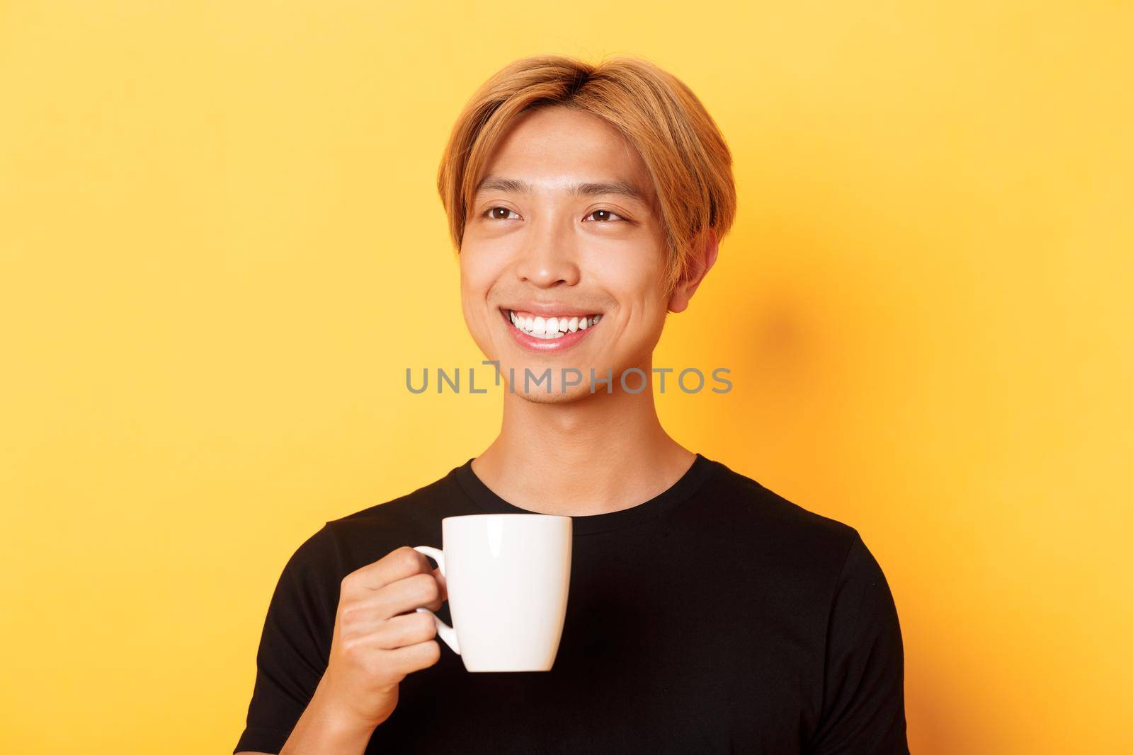 Close-up of happy handsome young asian guy with blond hair, looking dreamy and smiling while drinking coffee or tea, standing over yellow background by Benzoix