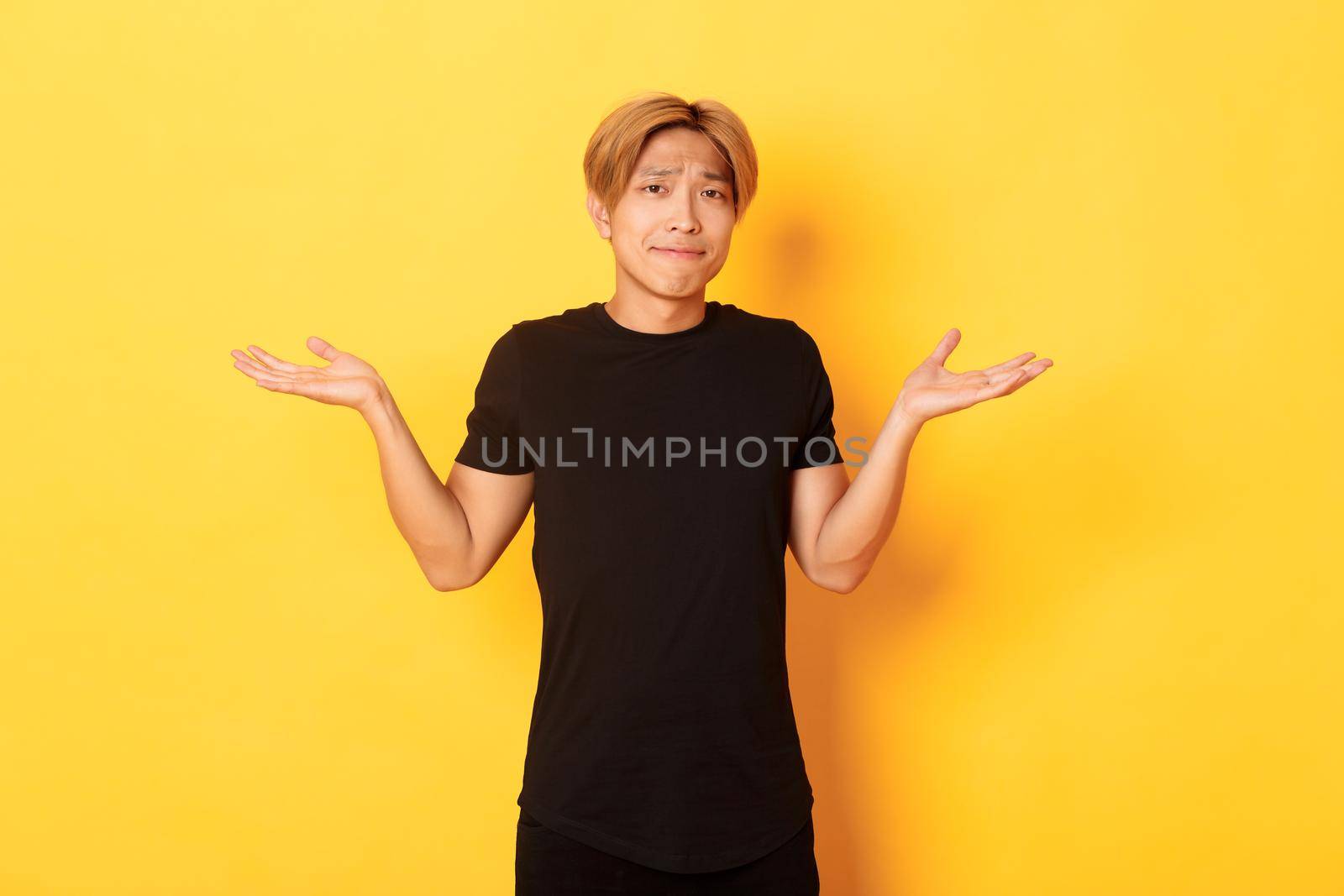 Portrait of confused and indecisive attractive korean guy, shrugging and pouting upset, standing yellow background.