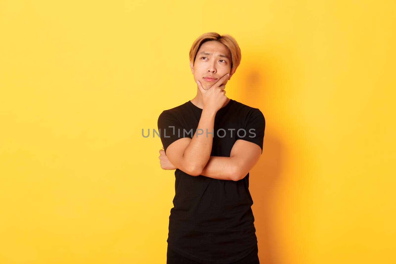 Portrait of thoughtful attractive guy in black t-shirt looking upper left corner. Young man pondering idea, standing yellow background.