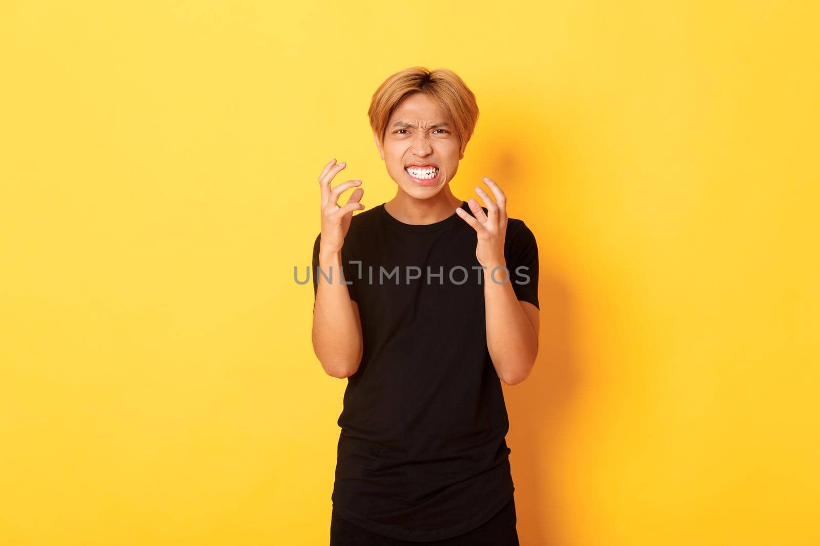 Portrait of angry and pissed-off asian blond guy, clenching hands furious and grimacing, standing over yellow background by Benzoix