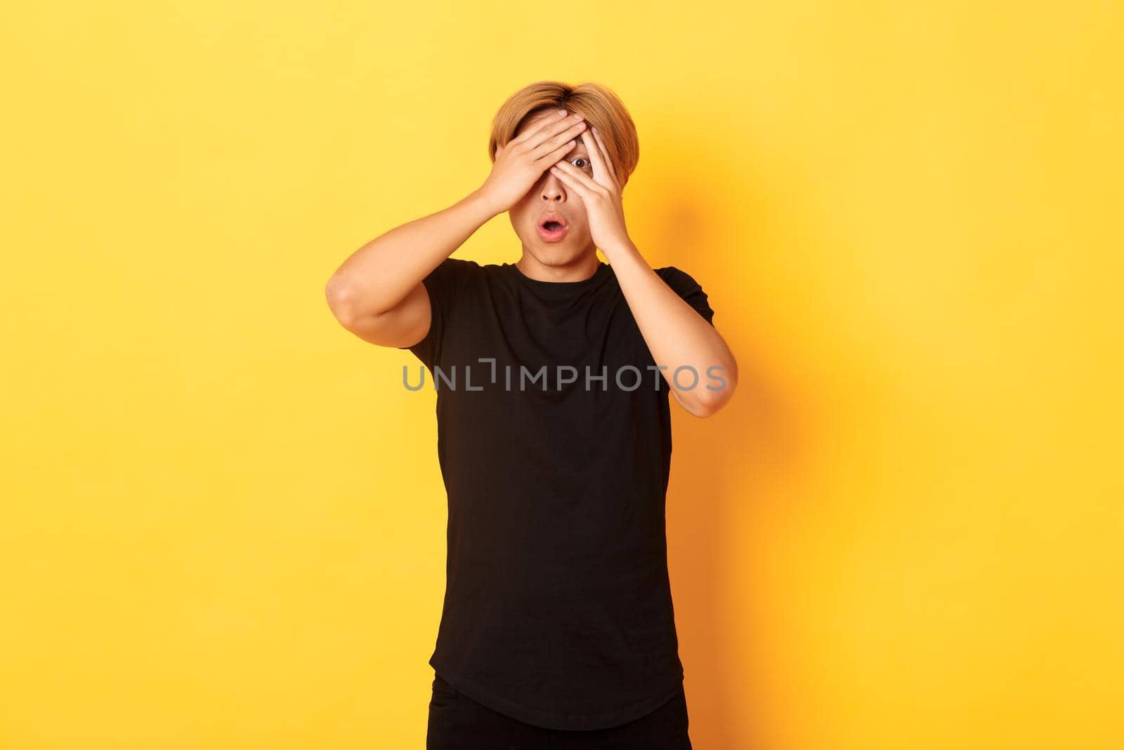 Portrait of embarrassed asian guy with blond hair, gasping startled and shut eyes, peeking through fingers, yellow background.