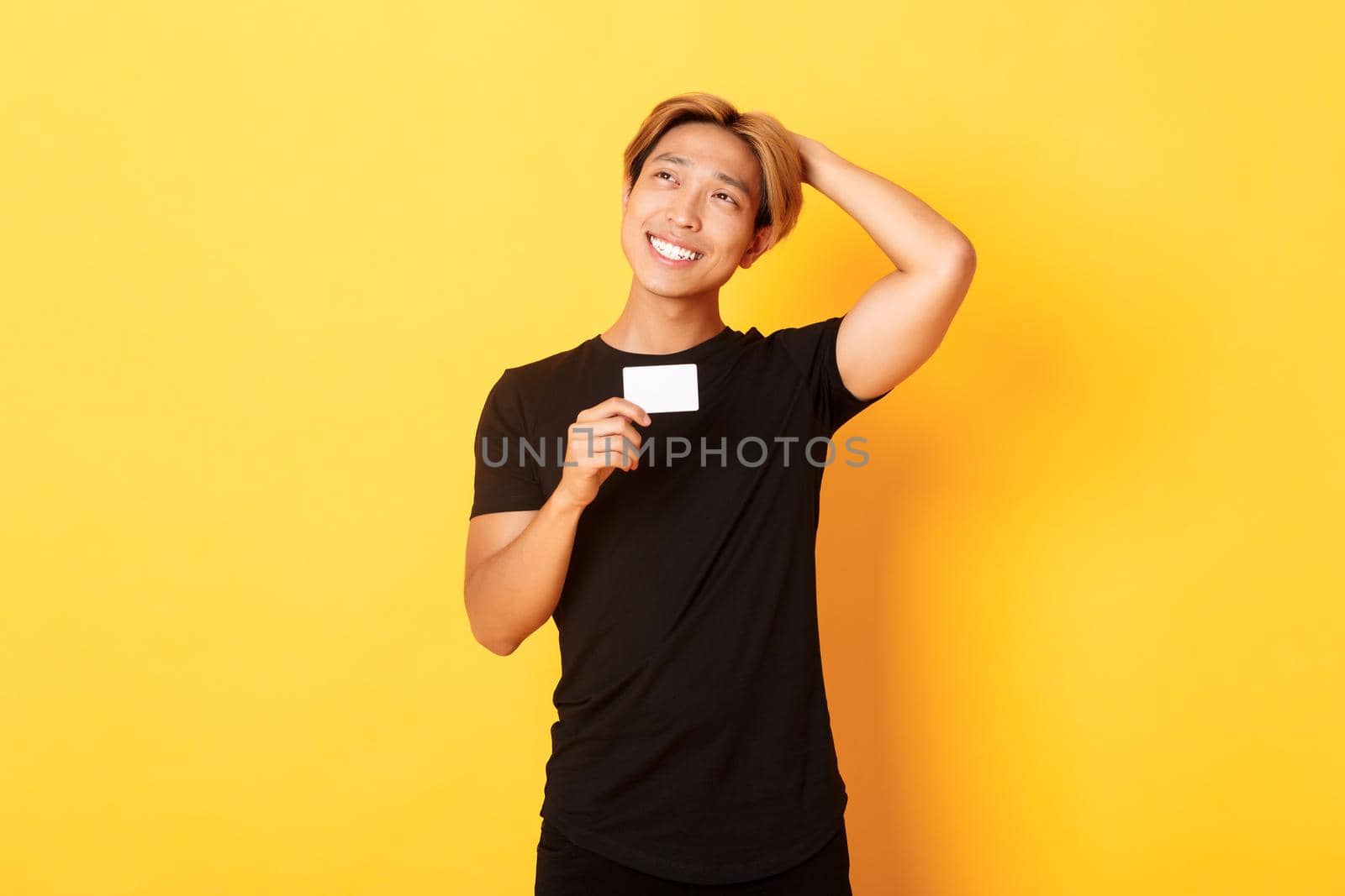Thoughtful smiling asian guy thinking while showing credit card, looking upper left corner dreamy, yellow background by Benzoix