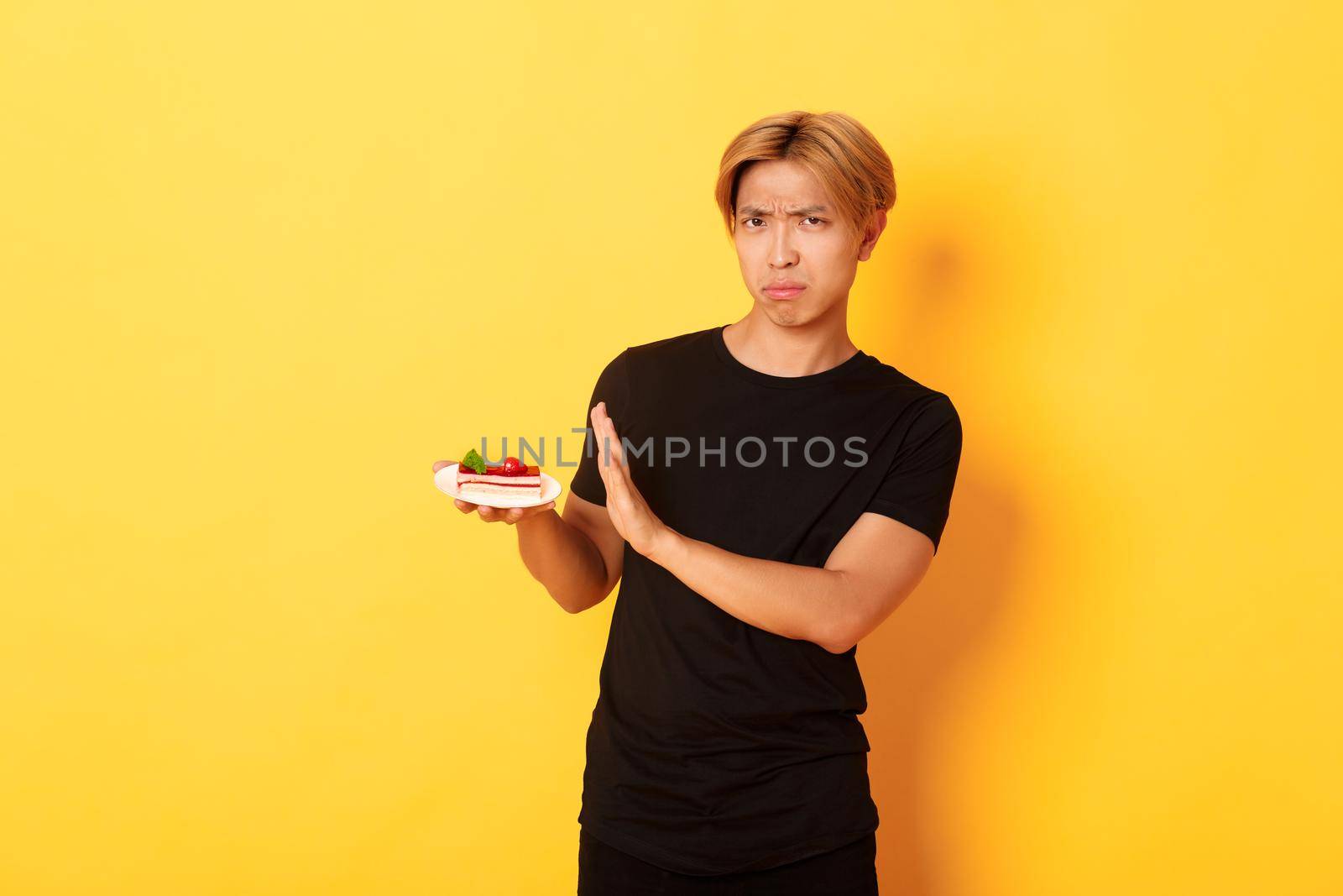 Portrait of disgusted and upset young asian guy refuse eating cake, looking displeased and showing rejection gesture, yellow background by Benzoix