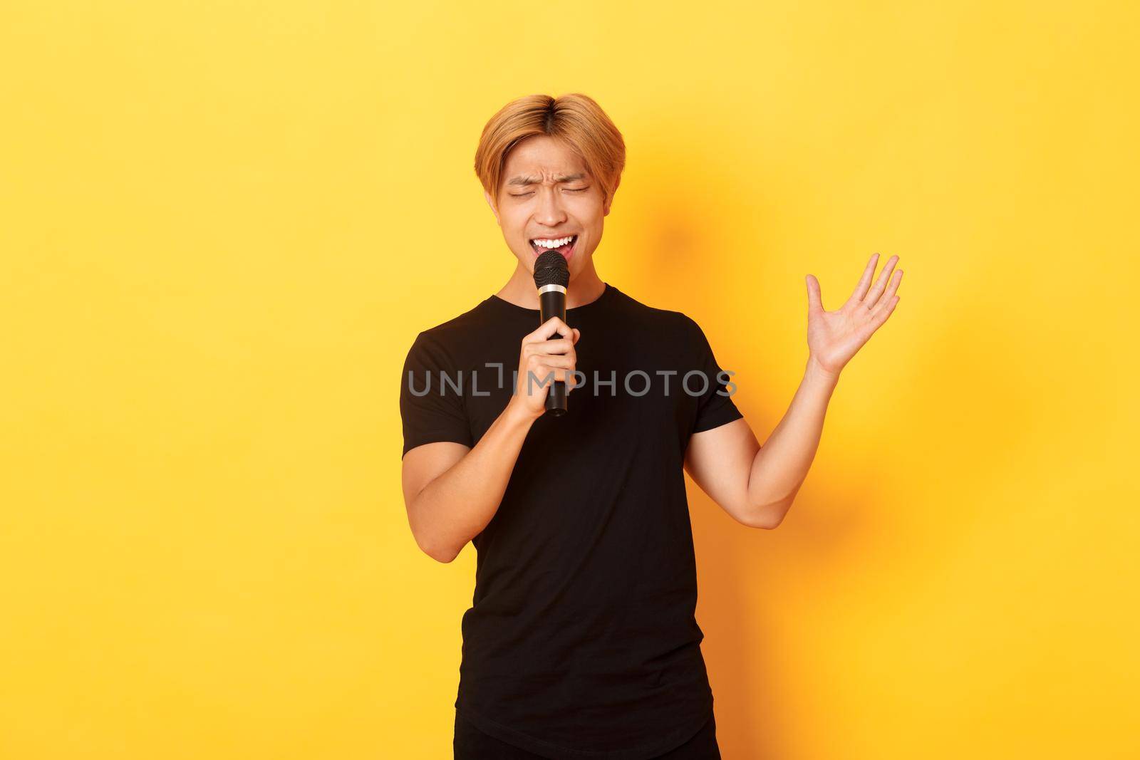 Handsome asian male singer, korean guy singing song at karaoke in microphone with passion, standing over yellow background.