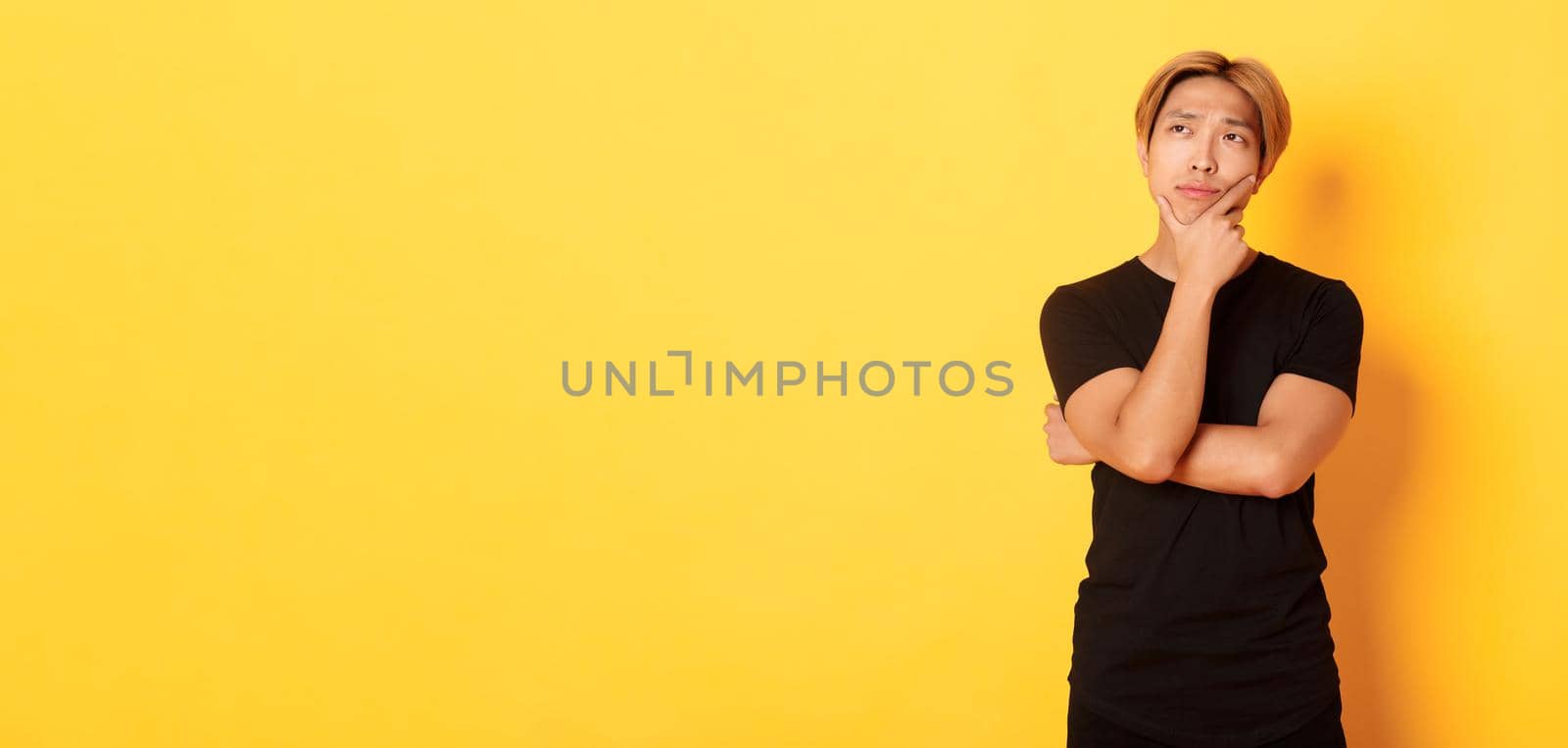 Portrait of thoughtful attractive guy in black t-shirt looking upper left corner. Young man pondering idea, standing yellow background by Benzoix