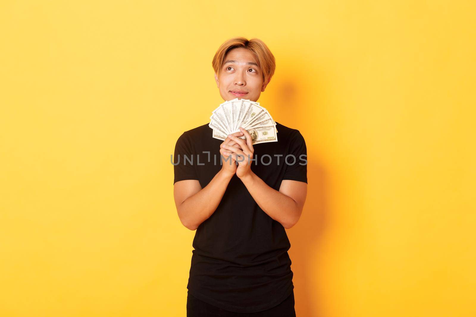 Portrait of dreamy handsome asian guy showing his savings and thinking, looking upper left corner, holding money, standing yellow background.