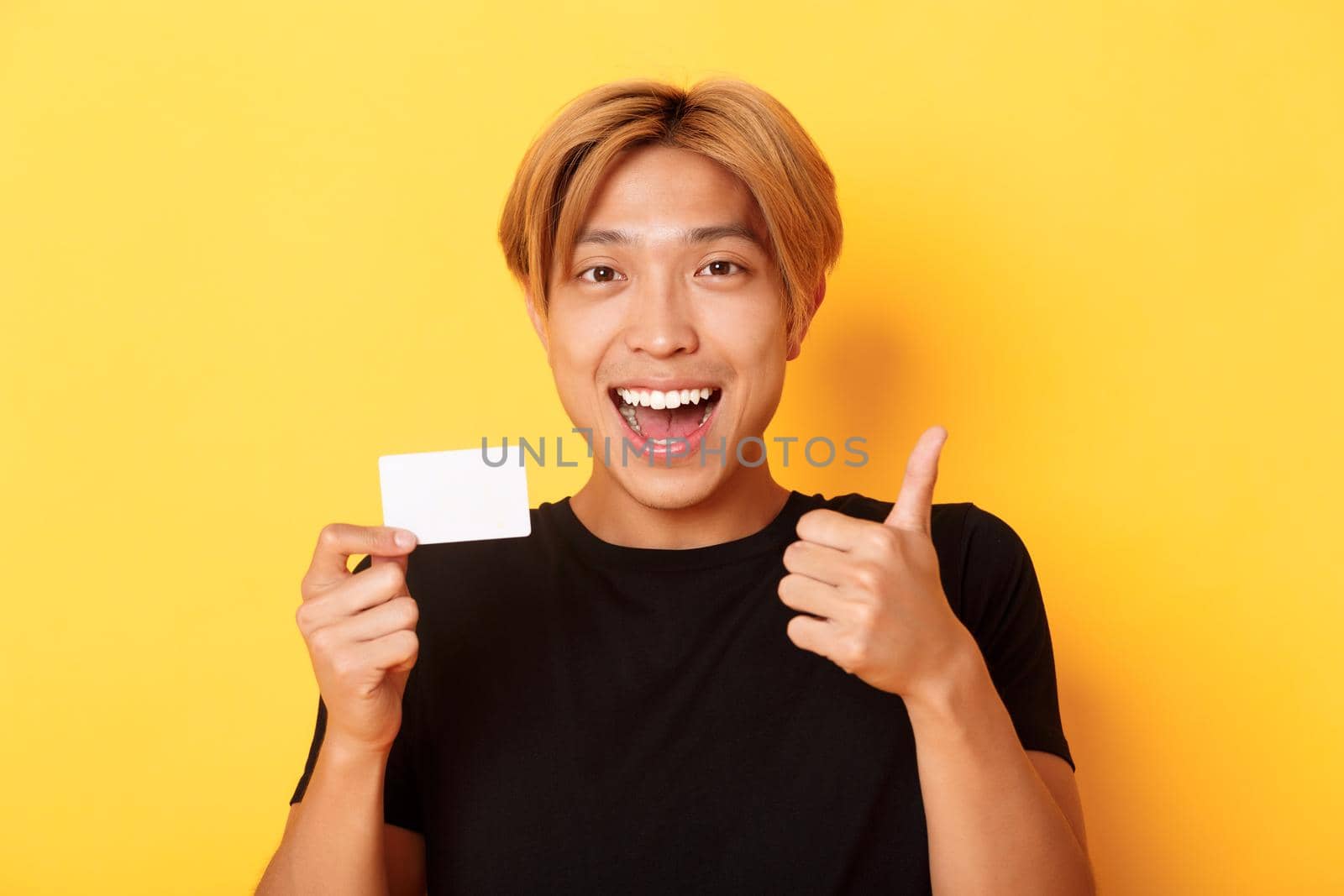 Close-up of happy and satisfied asian handsome guy, showing credit card and thumbs-up in approval, smiling amazed, standing yellow background.