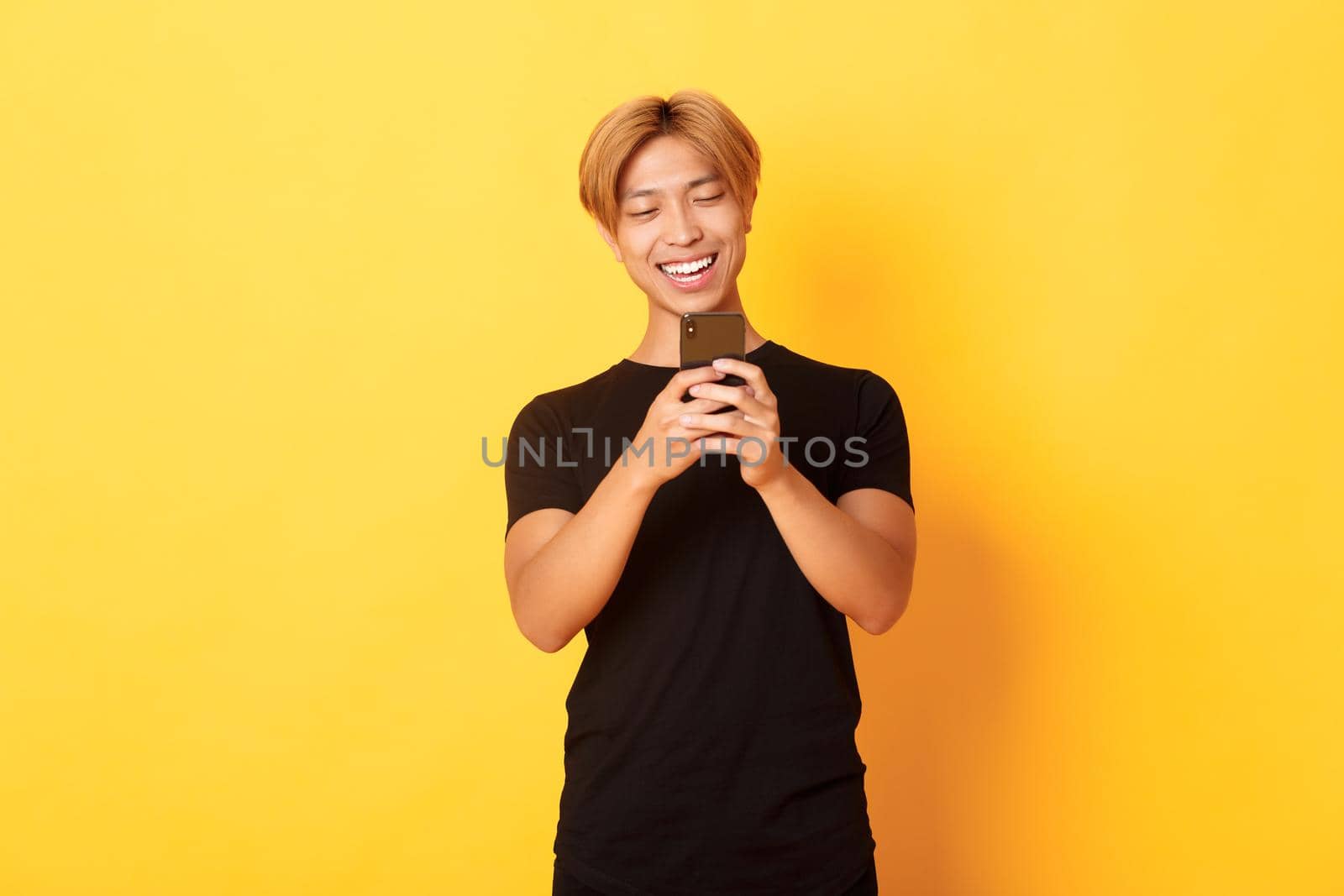 Portrait of handsome stylish asian guy with blond hair, using mobile phone and smiling, messaging in smartphone app, yellow background.