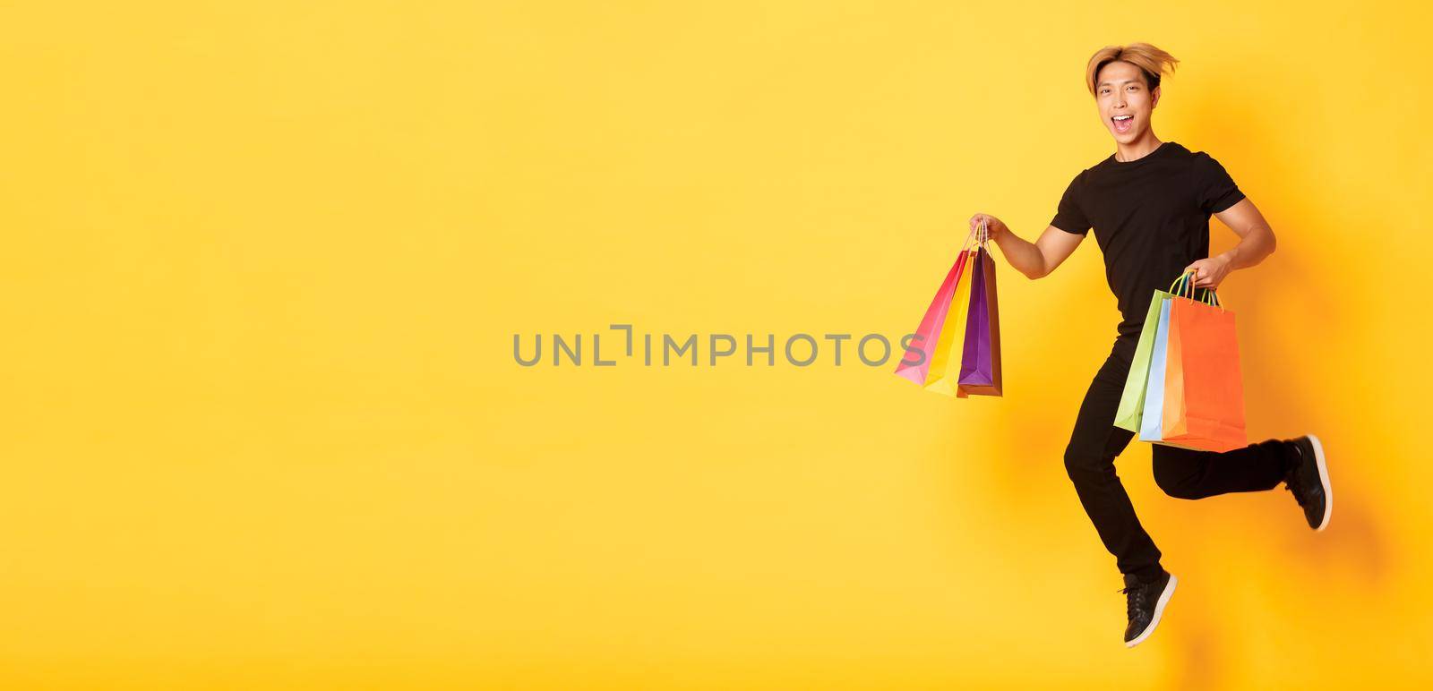 Full-length of happy attractive asian man jumping from happiness and carry shopping bags, standing yellow background.