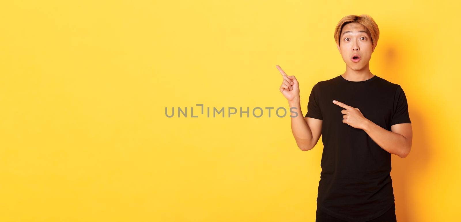 Portrait of impressed and excited handsome asian guy in black t-shirt, react to your logo, pointing fingers upper left corner.