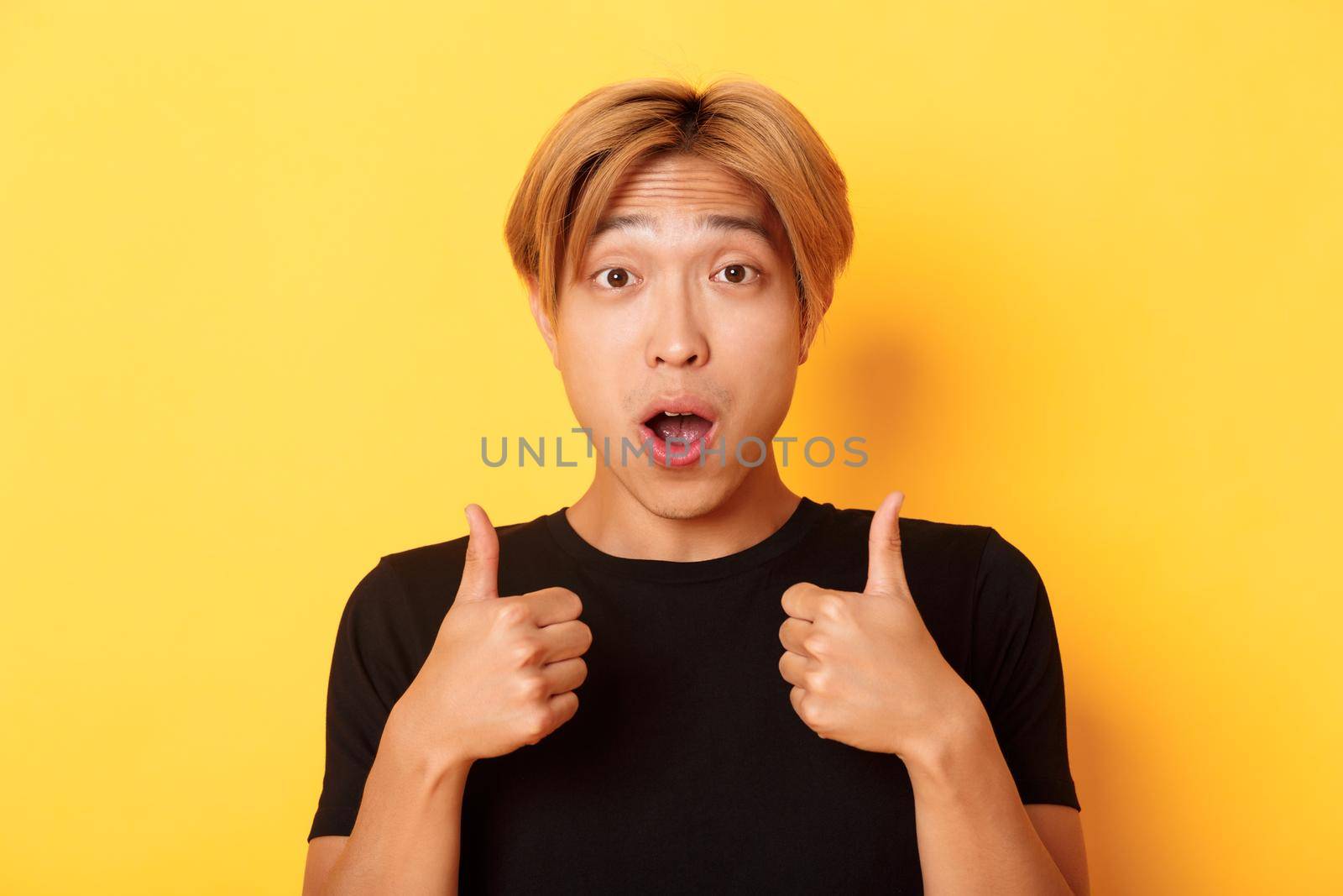 Close-up of impressed and excited handsome asian guy showing thumbs-up and looking amazed, standing over yellow background.