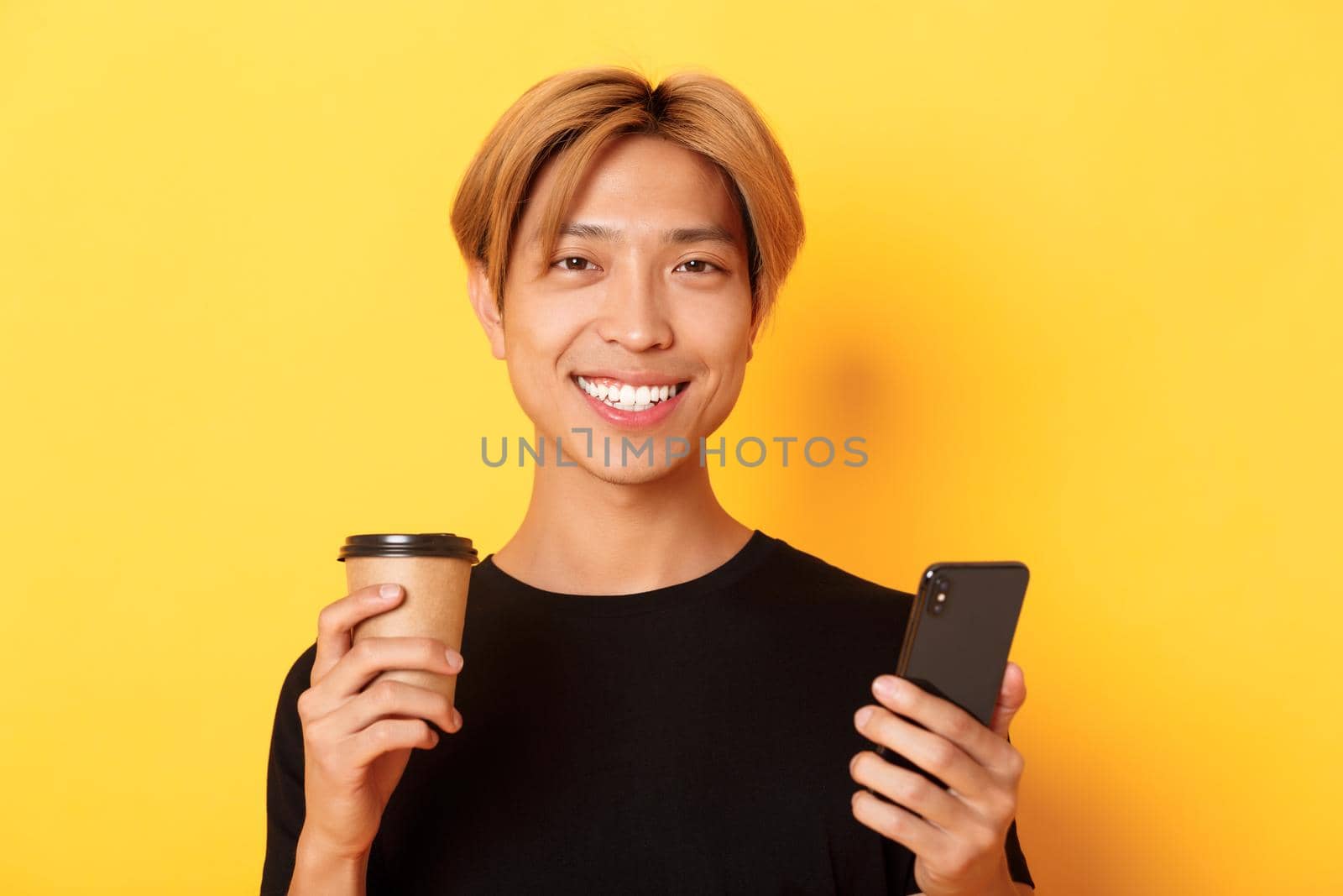 Close-up of handsome young asian guy smiling happy, using smartphone and drinking takeaway coffee, standing over yellow background.