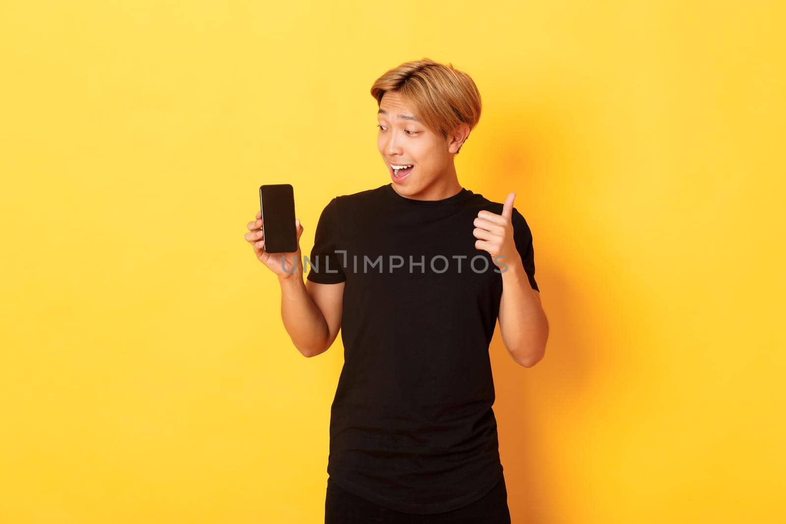 Portrait of satisfied asian guy looking at smartphone screen and showing thumbs-up in approval, standing yellow background.