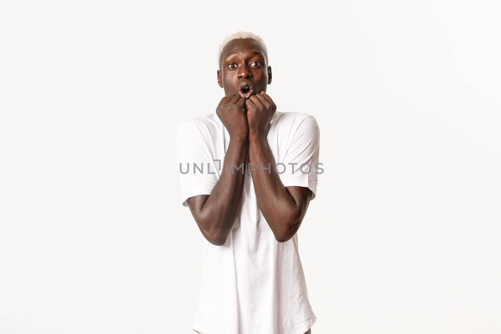 Portrait of amazed and fascinated african-american blond man, gasping excited, holding hands pressed to face, looking at camera thrilled, standing white background.