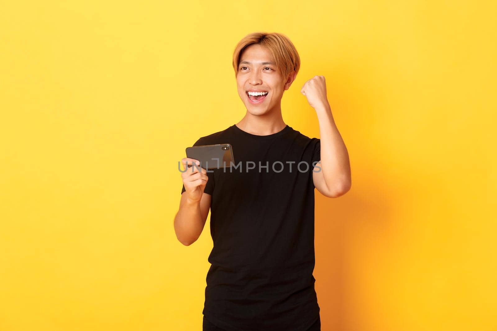 Portrait of stylish handsome asian guy playing smartphone game, rejoicing from winning, standing yellow background.