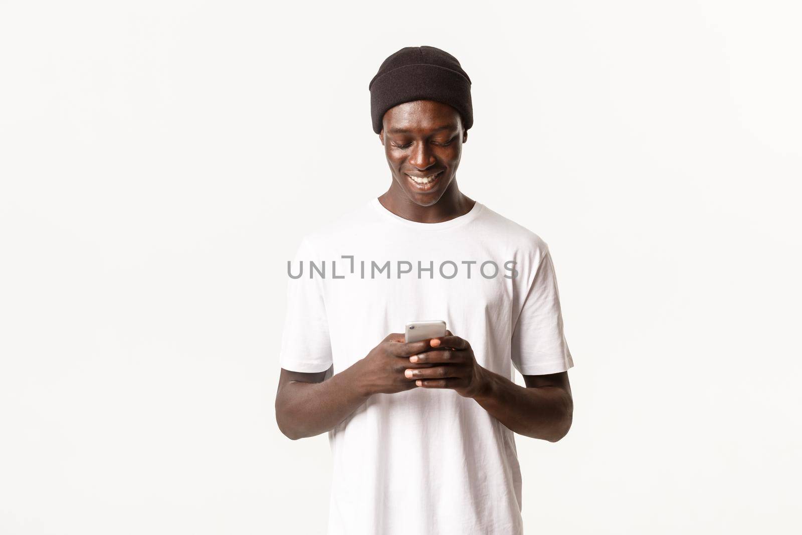 Portrait of handsome african-american young man in beanie using mobile phone, messaging and smiling at smartphone screen, white background by Benzoix