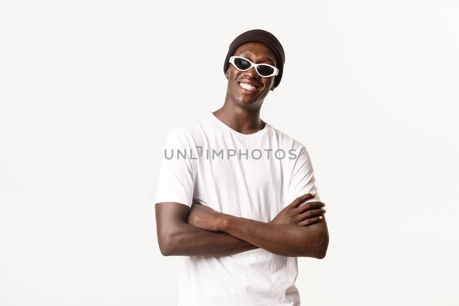 Portrait of sassy and stylish african-american guy in beanie, sunglasses, standing pleased with arms crossed and smiling happy at camera, white background by Benzoix
