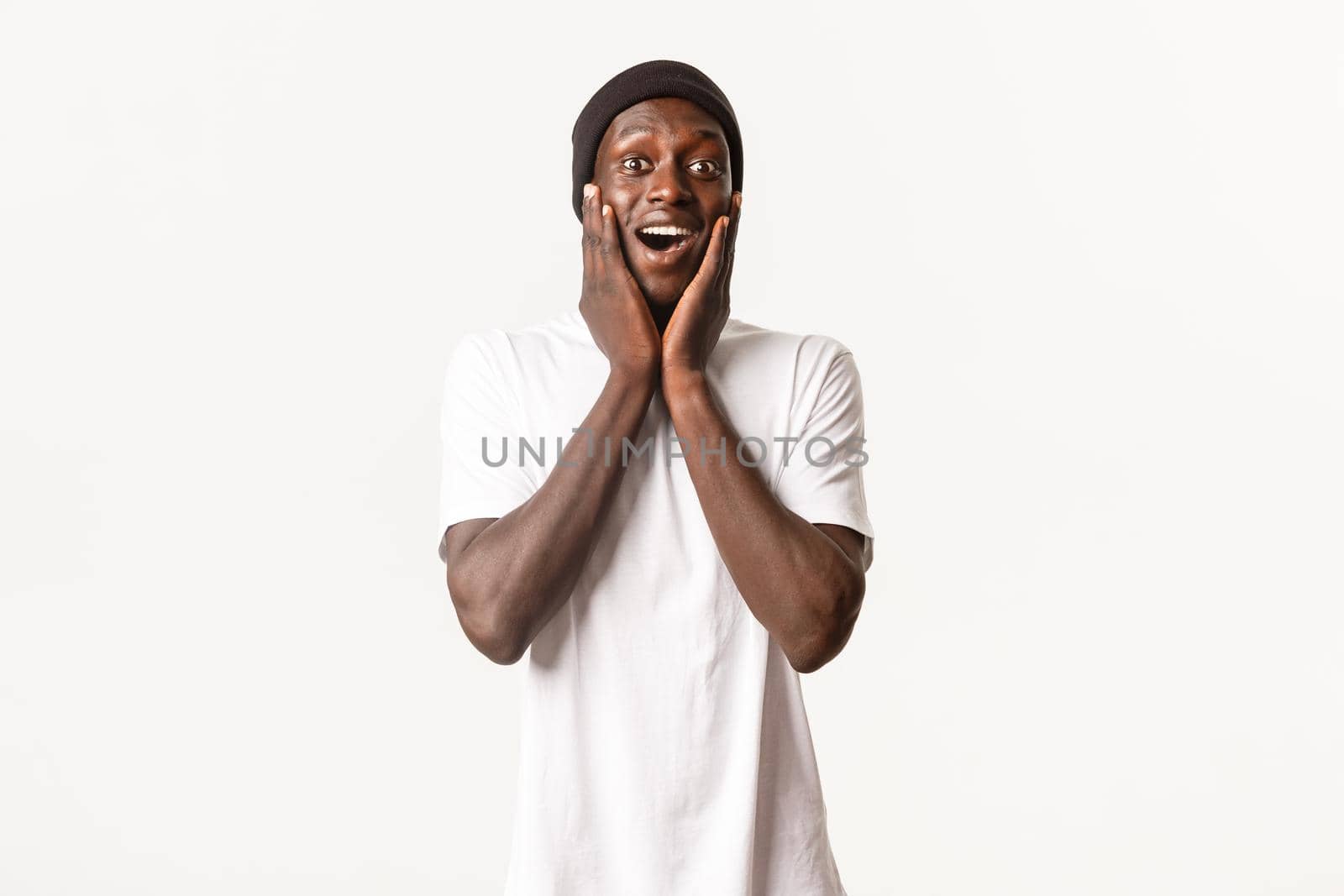 Portrait of happy surprised young african-american guy, looking amazed and cheerful, gasping fascinated, holding hands on cheeks, white background.
