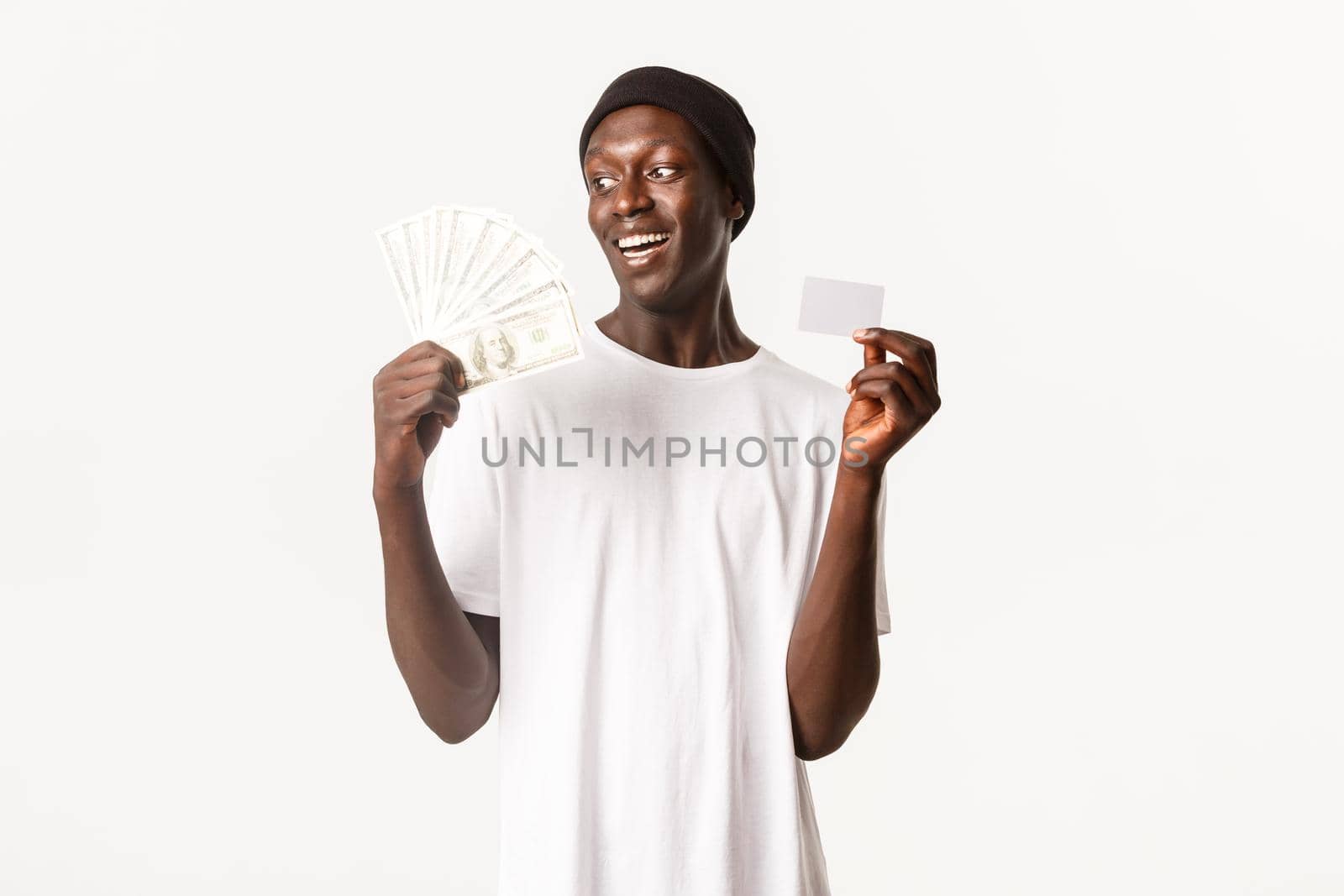 Portrait of lucky, excited african-american young guy looking happy at money, holding credit card, white background.