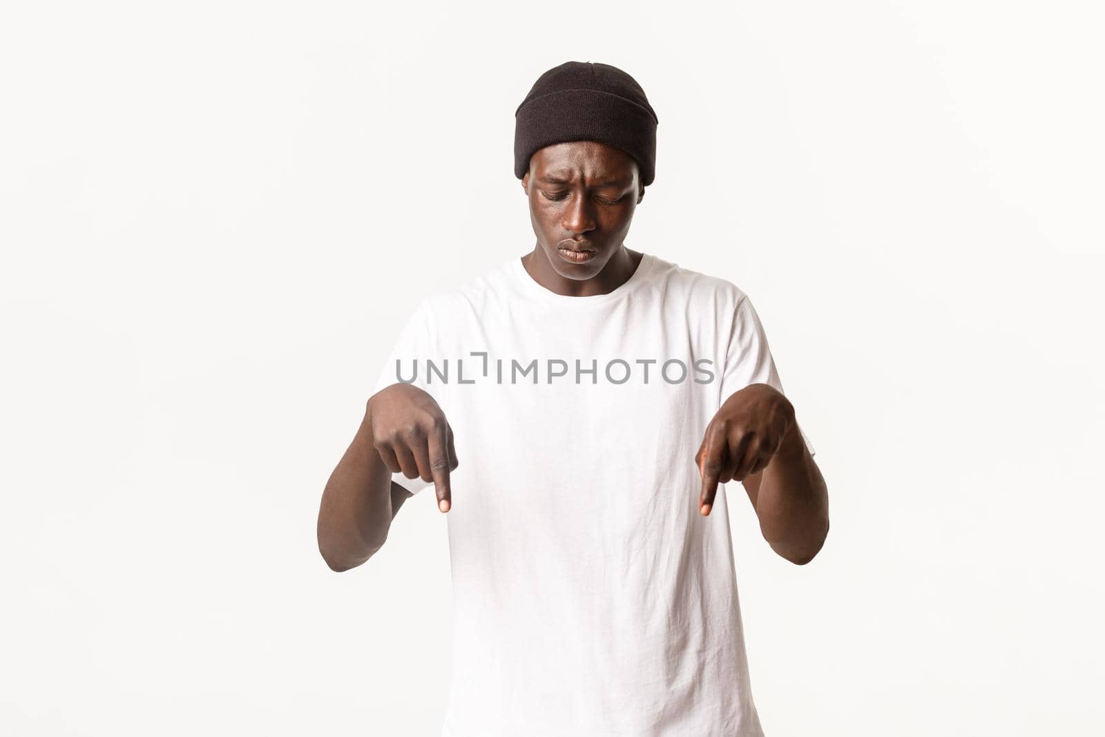 Portrait of confused african-american guy in beanie, looking perplexed and pointing fingers down, standing puzzled over white background by Benzoix